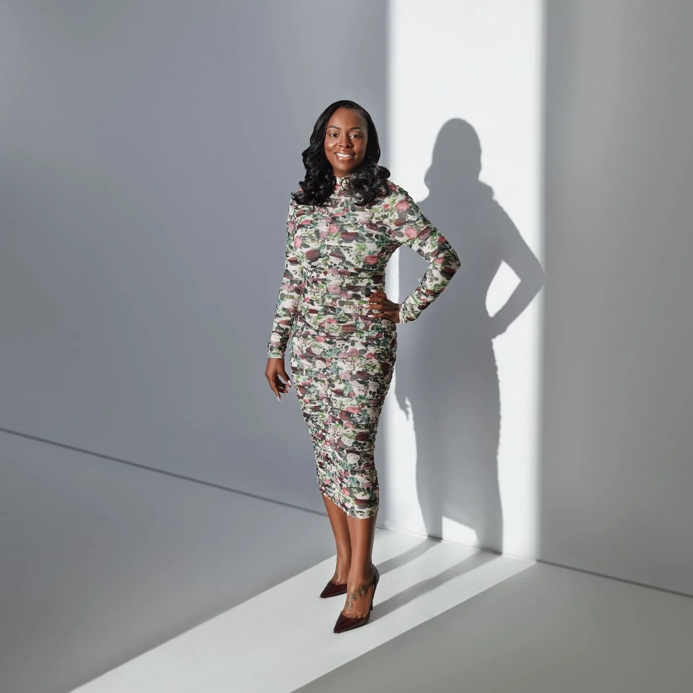 A woman in a floral dress and high heels standing against a plain wall with a shadow cast behind her.