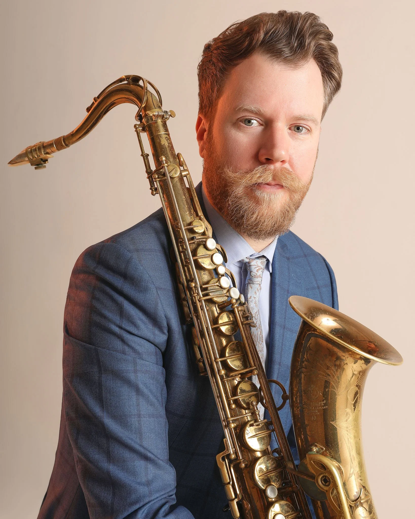 A man with a beard and mustache wearing a blue suit jacket and tie, holding a brass saxophone over his shoulder, looking at the camera against a plain beige background.