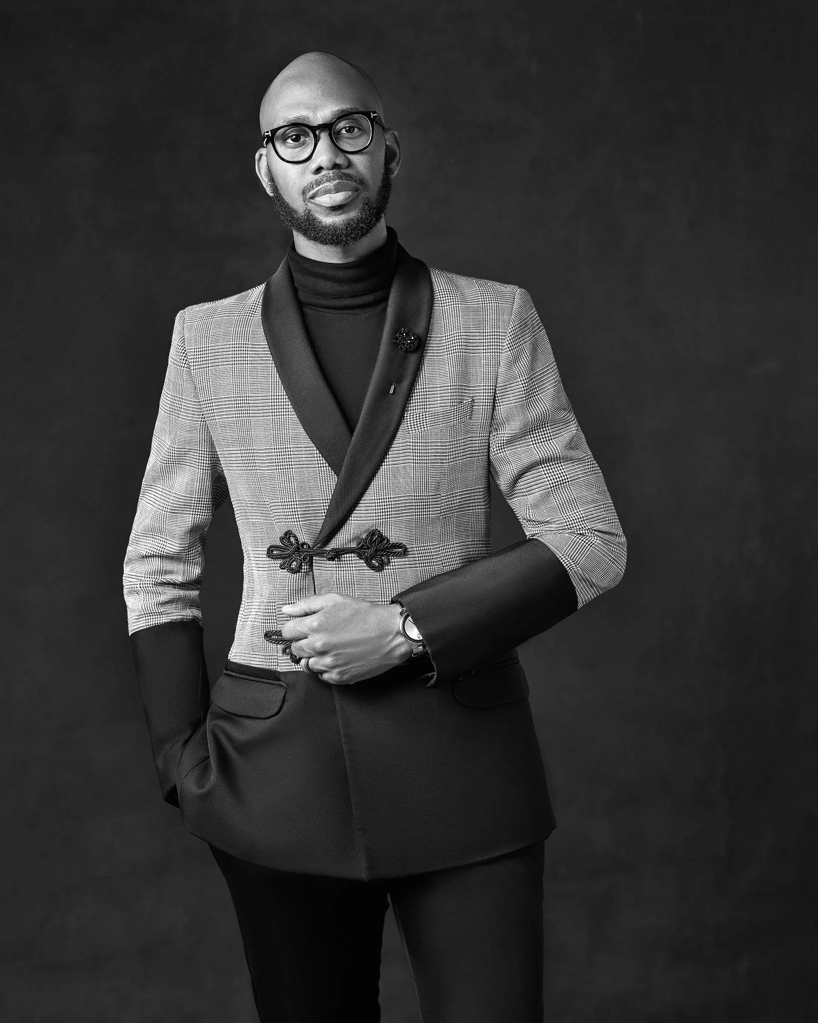 Portrait of a man with glasses dressed in a stylish blazer and turtleneck posing against a dark background.