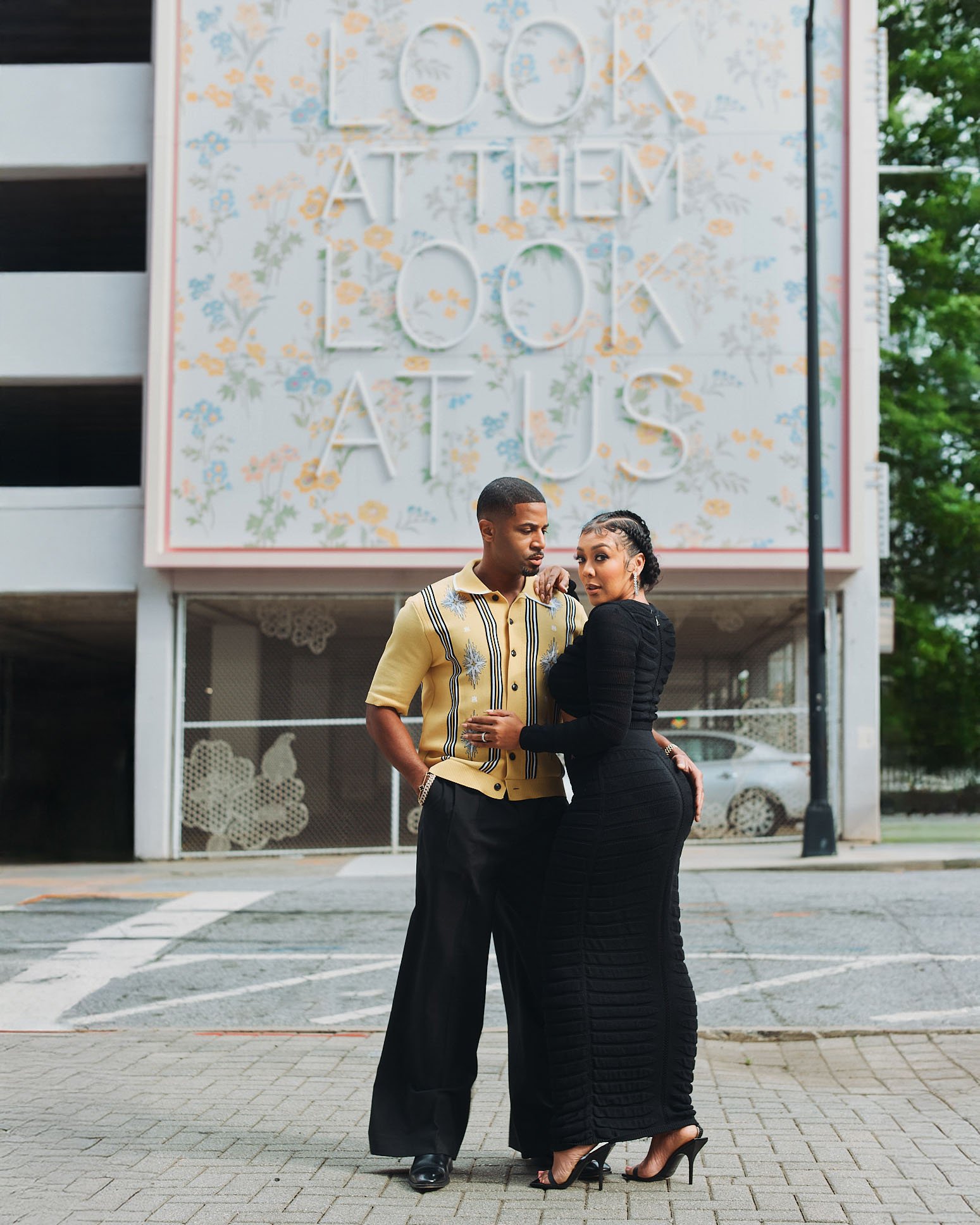A man and woman standing closely together outdoors in front of a parking garage and a large floral sign that reads 'LOOK AT THEM LOOK AT US.'