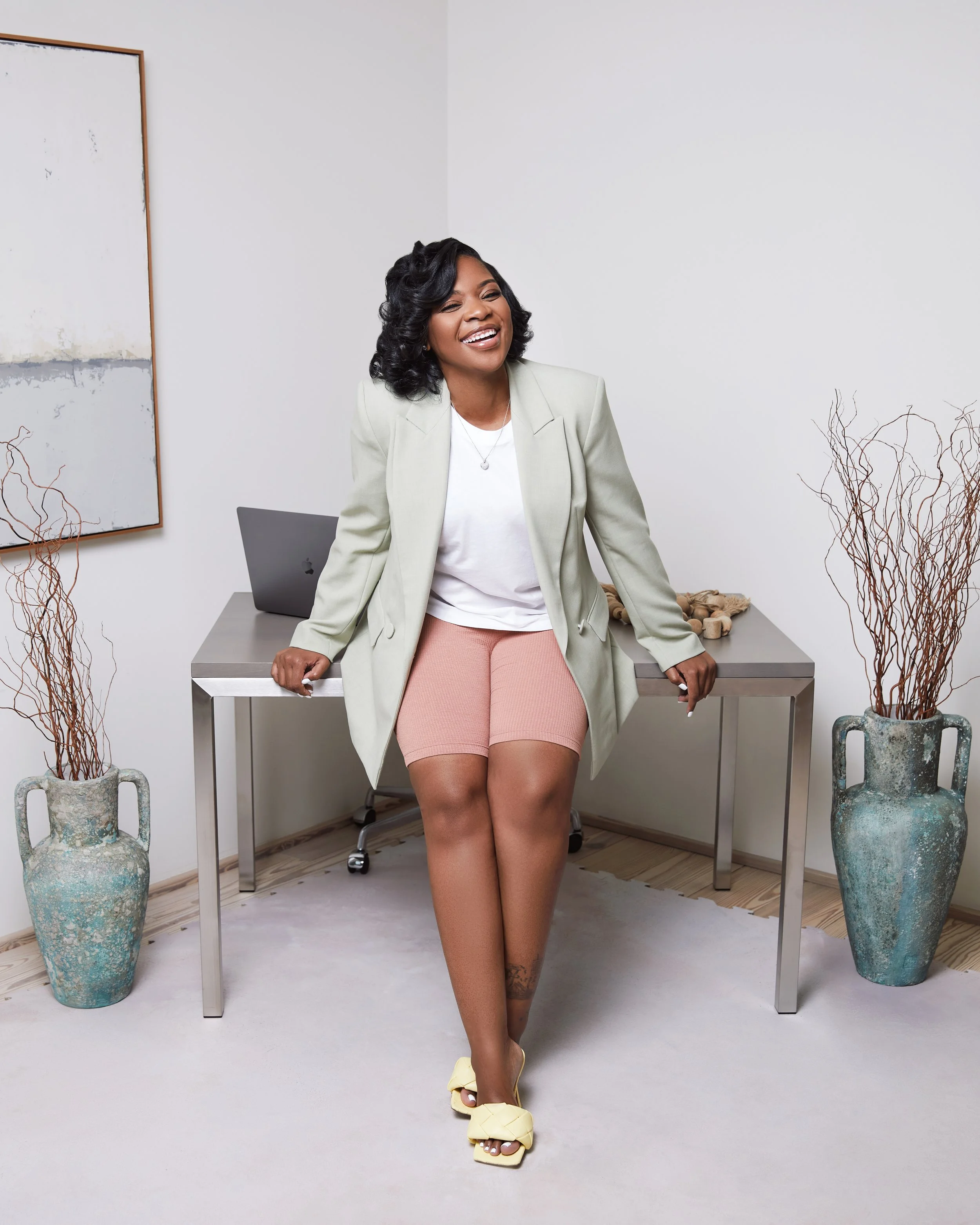 A woman sitting on the edge of a desk in an office, smiling, wearing a light blazer, white shirt, peach shorts, yellow sandals, and sheer stockings. Two large vases with dried branches are on either side of the desk.