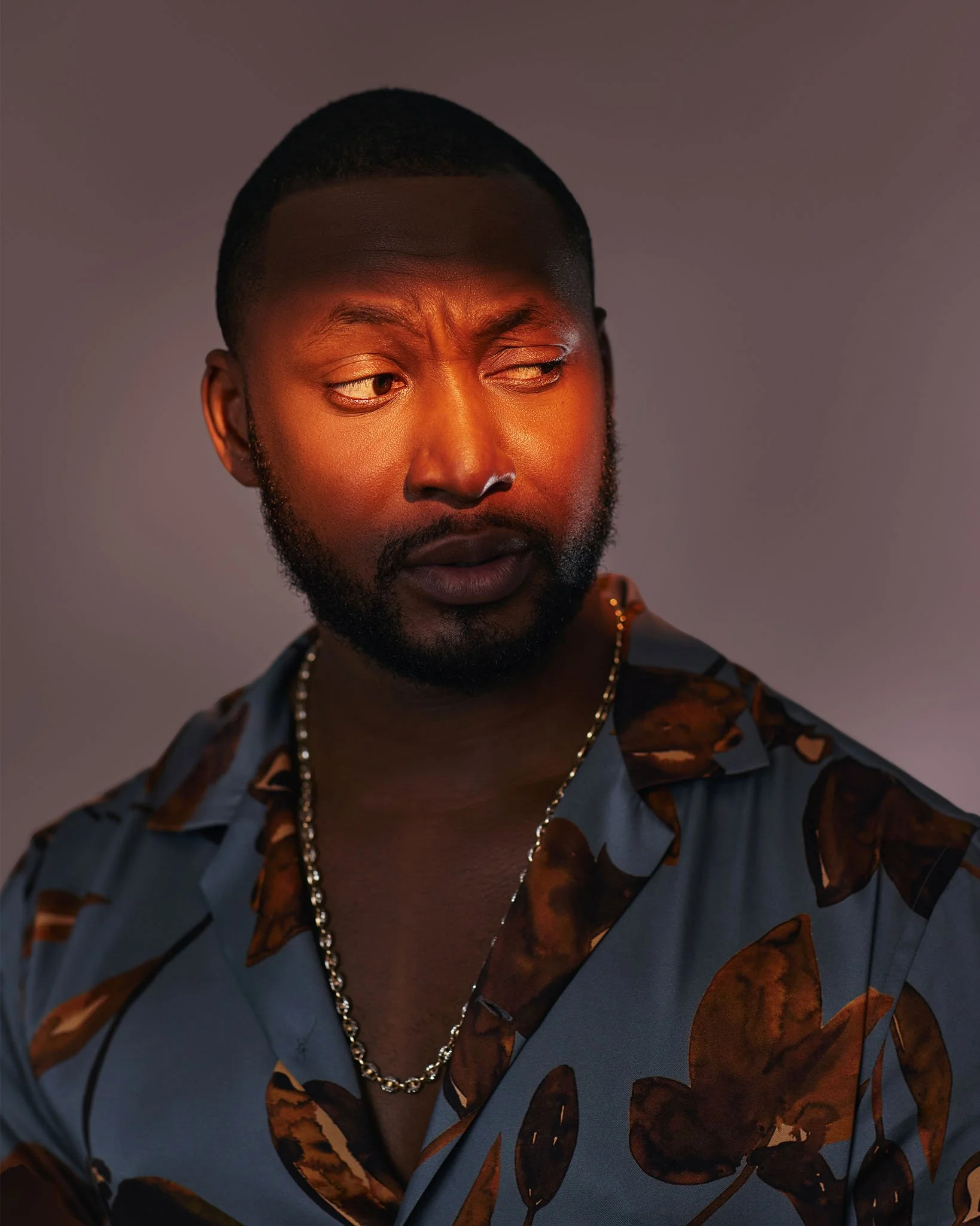 A young man with a beard and short hair looking to his left, wearing a patterned shirt with a chain necklace, against a neutral background.
