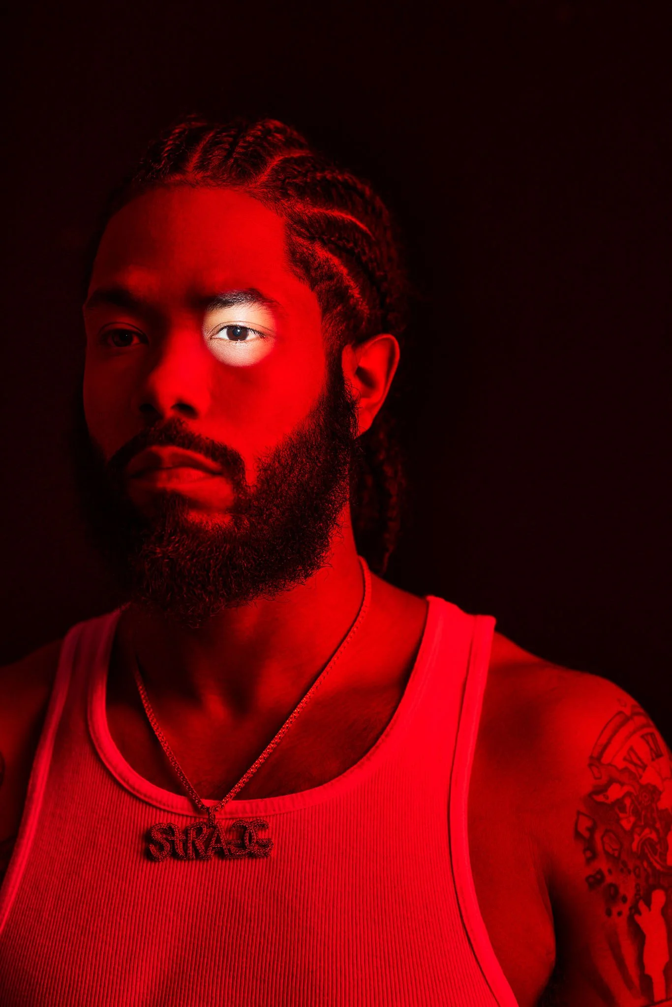 A man with braided hair and a beard wearing a white tank top and a necklace with the word 'STRANGE' written on it, illuminated with red lighting against a dark background.