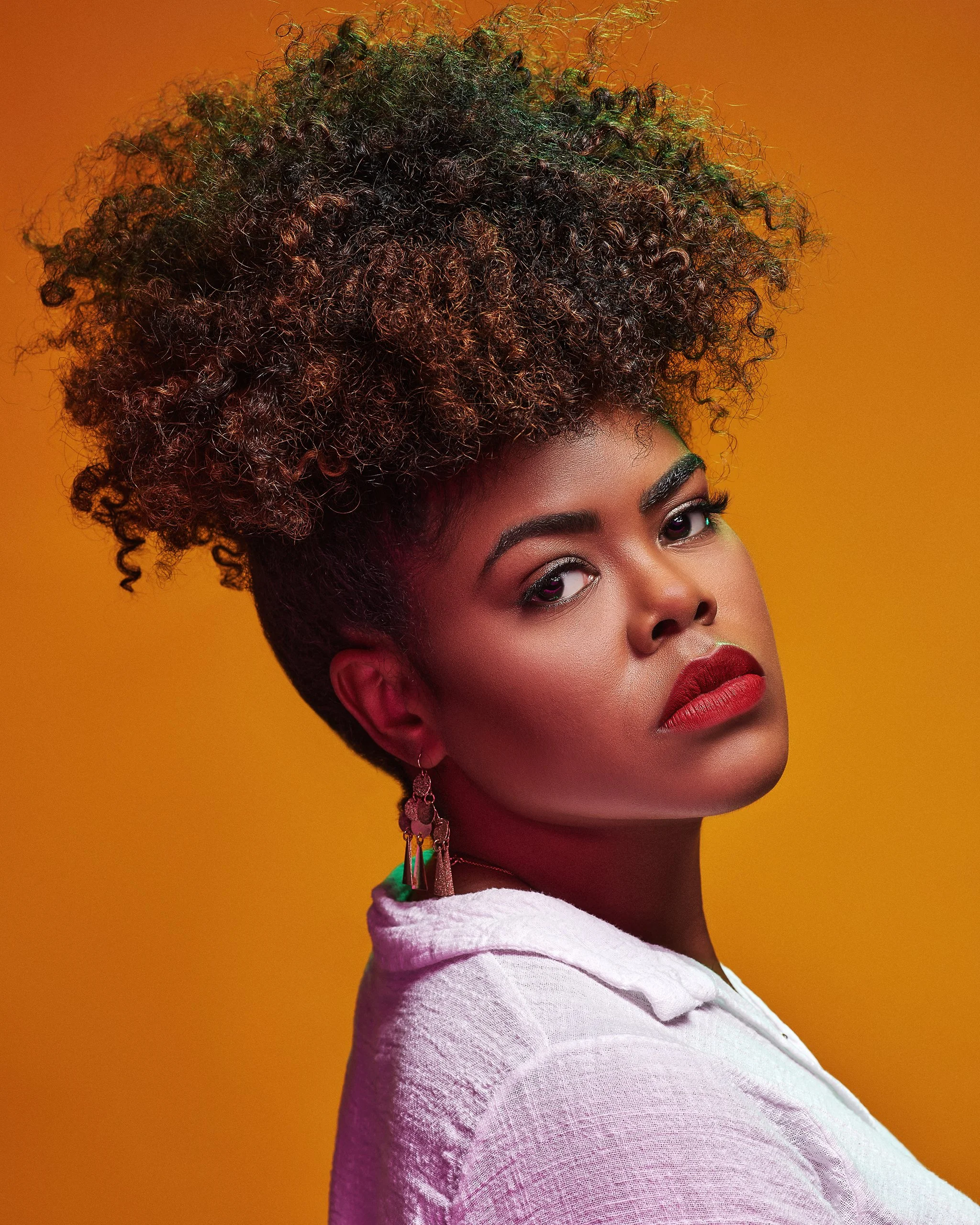 A woman with a modern hairstyle of voluminous tight curls styled up, wearing a white textured top and colorful earrings, posing against a yellow-orange background.