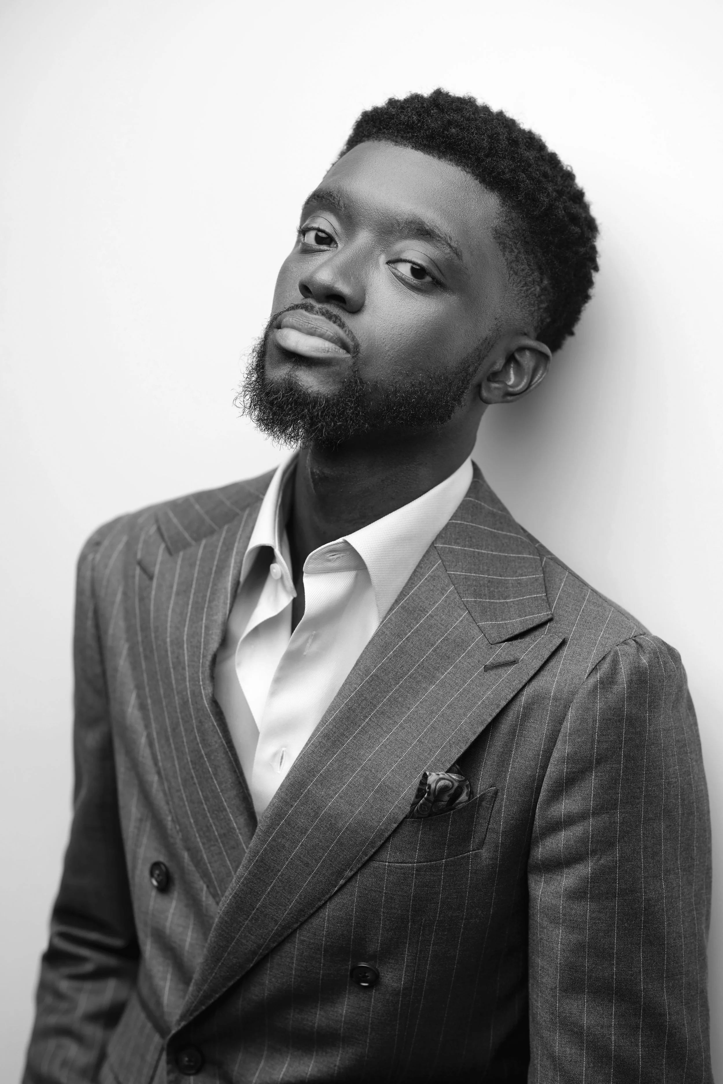 Black and white portrait of a young man in a pinstripe suit with a pocket square and an open-collared shirt, standing against a plain background, looking confidently at the camera.