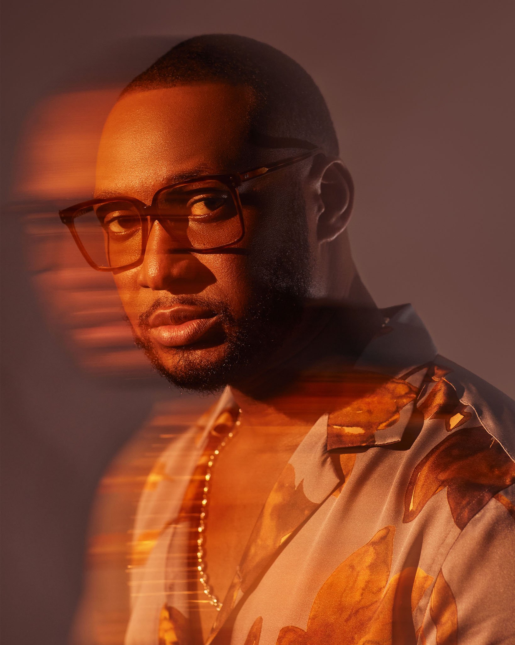 A portrait of a man with glasses and a beard, wearing a patterned shirt and a gold necklace, with a blurred motion effect and warm lighting.