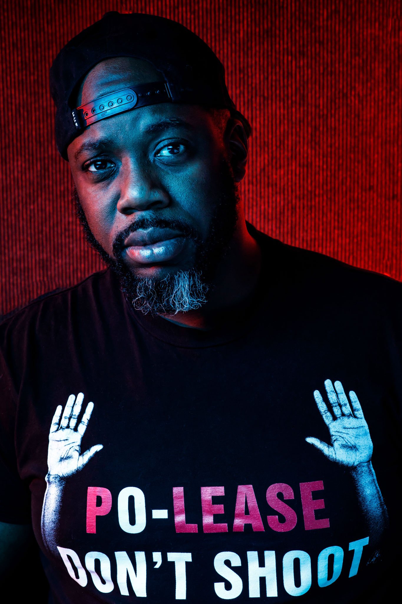 A man wearing a backwards black cap and a black T-shirt with white and pink text that reads 'PO-LEASE DON'T SHOOT'. The background is a red textured wall, and the man has a serious expression with a gray beard.