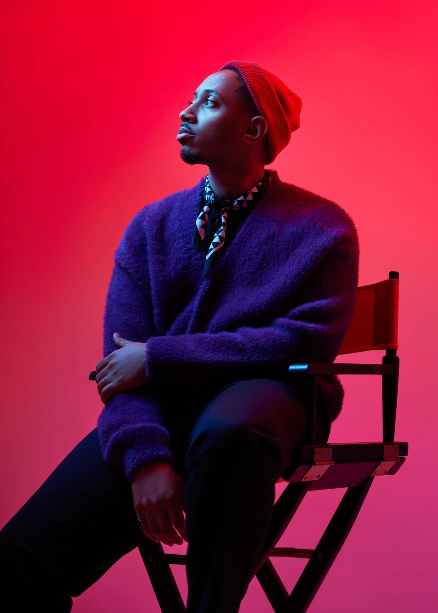 A man with short hair and a beard, wearing a red beanie, patterned neck scarf, and a purple fuzzy sweater, sitting on a black folding chair against a pinkish-red background, looking to the left.