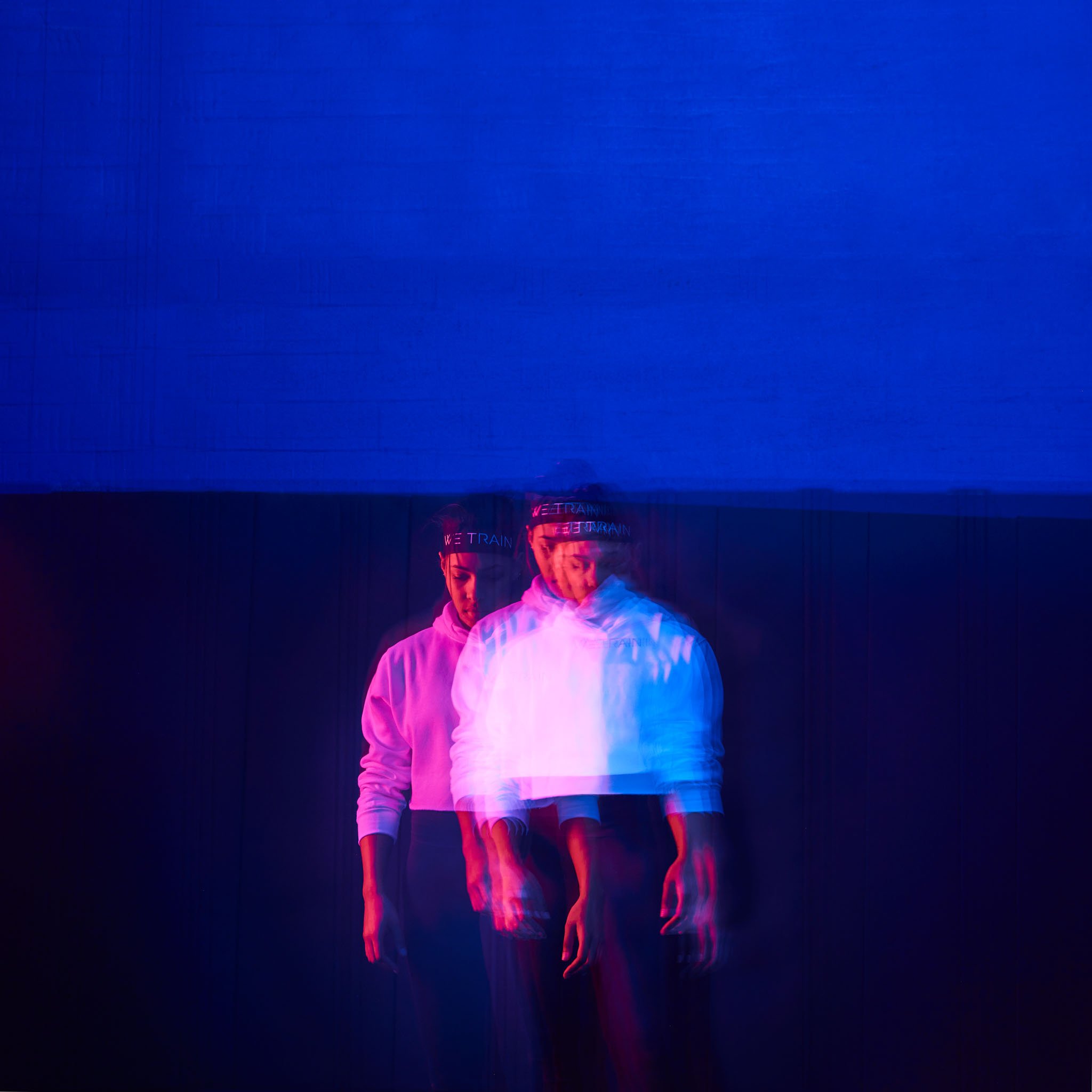 A person wearing a white hoodie and a black headband with 'WE TRAIN' written on it, walking in a space with a blue and pink neon glow, with a motion blur effect showing multiple overlapping images of the person.