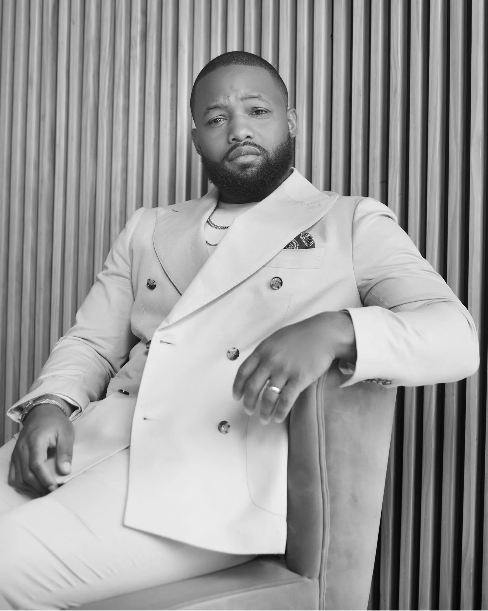 A man with a beard and short hair, wearing a light-colored double-breasted suit with a pocket square, sitting on a chair against a vertical wood panel background in black and white.