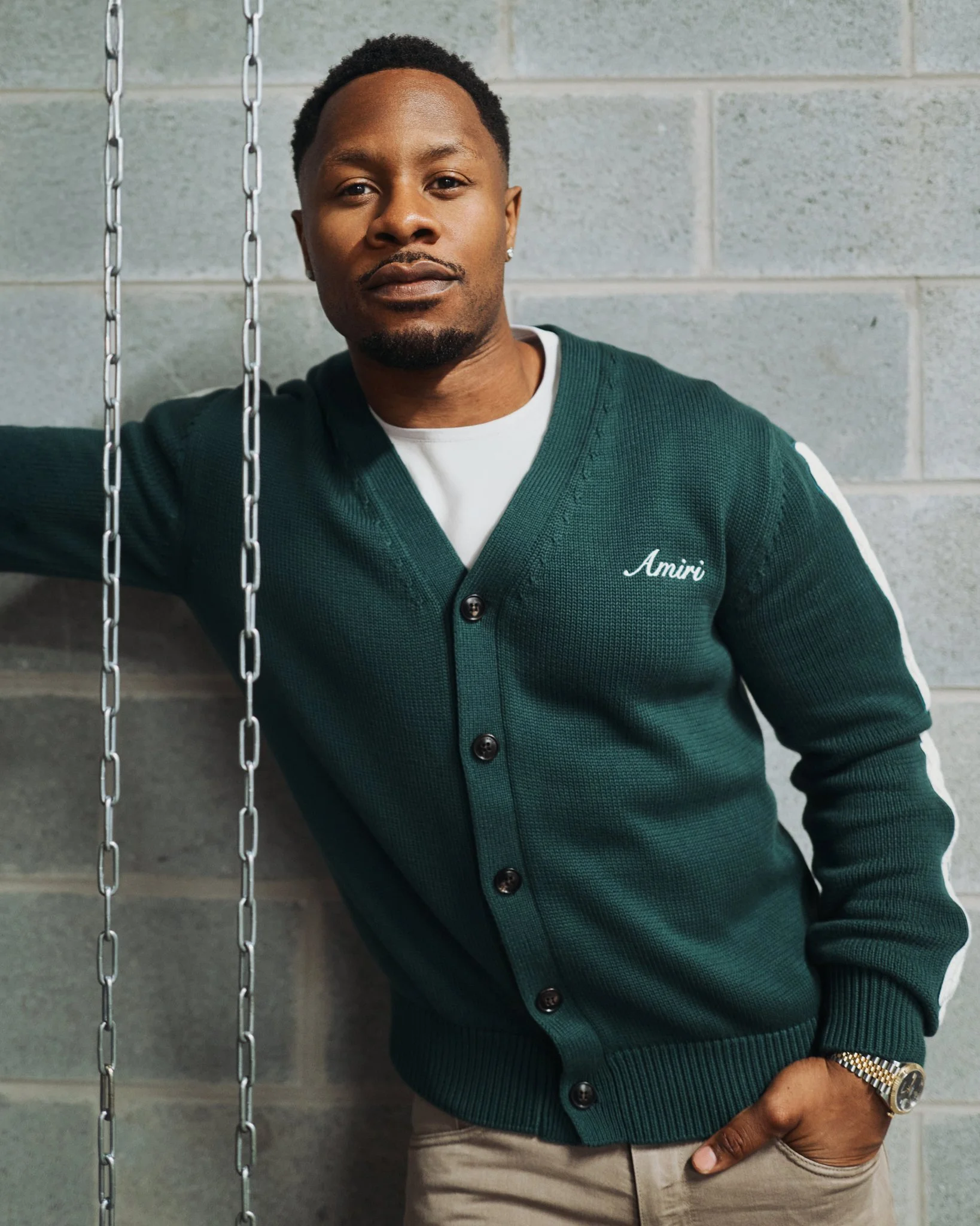 Chido Awokocha , wearing a green cardigan with the name 'Amiri' embroidered on it, standing against a gray cinder block wall, looking confidently at the camera.