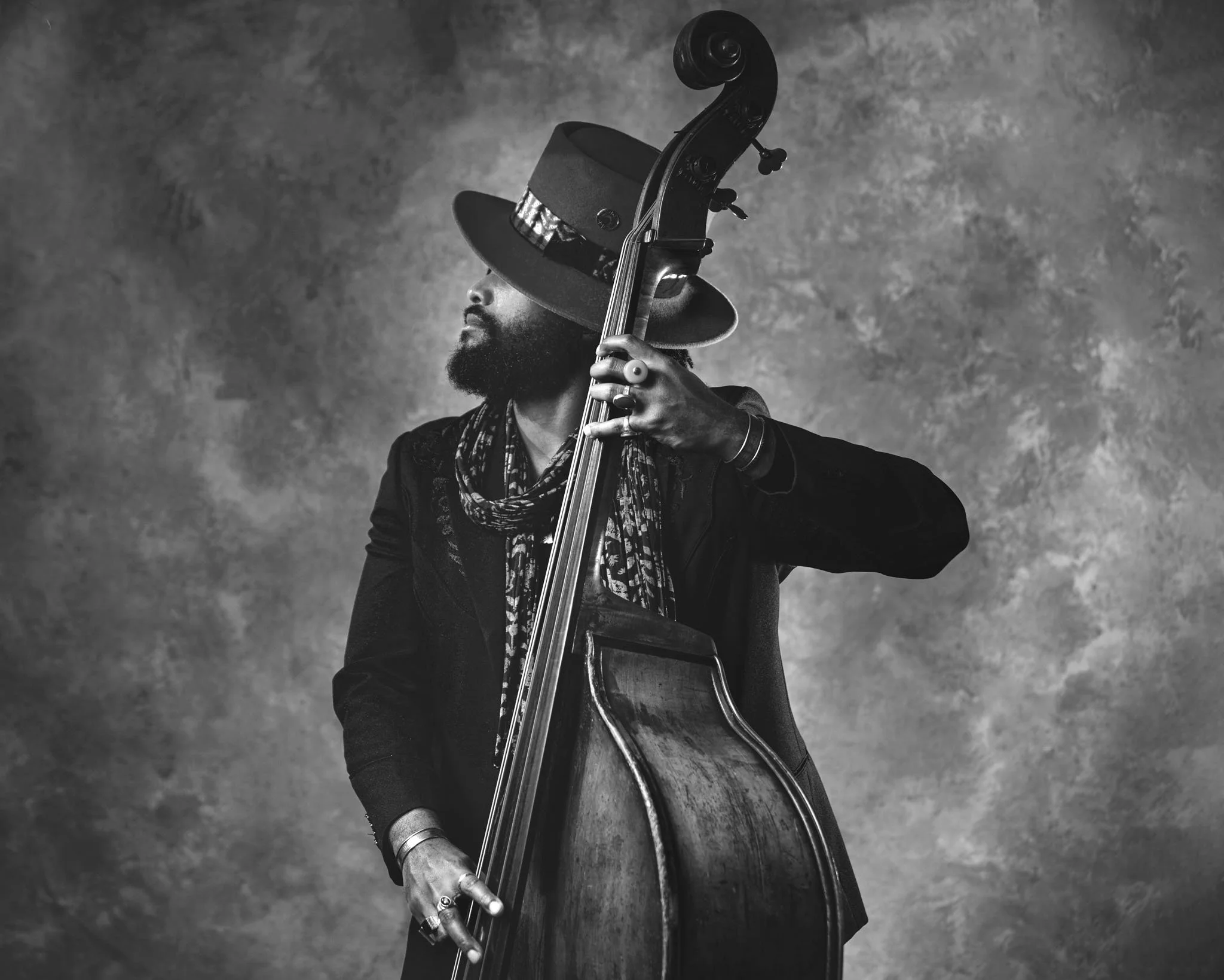 A man with a beard wearing a large Hat, scarf, and dark clothing plays a double bass against a textured background.