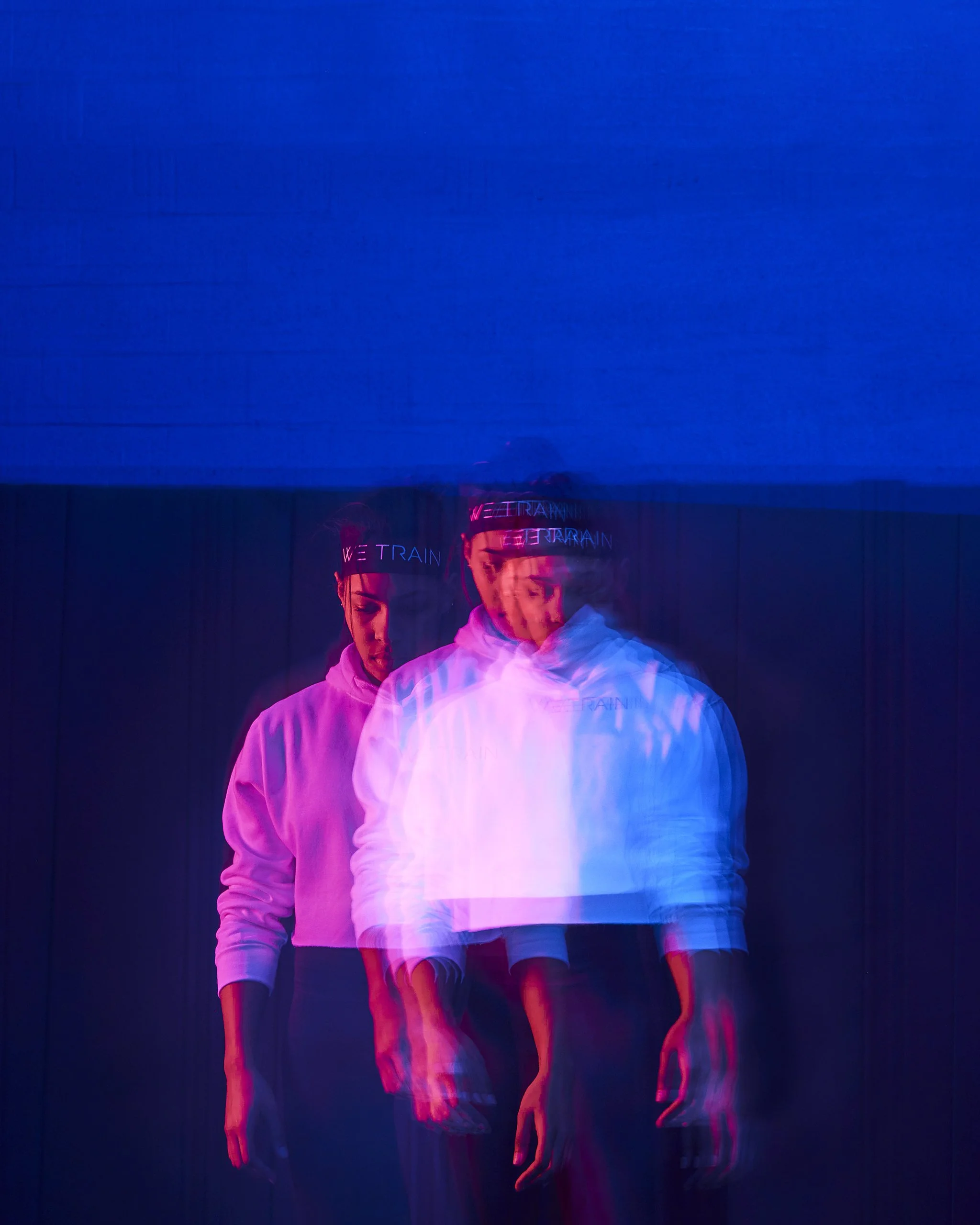 Multiple overlapping images of a woman in a white hoodie and a headband with neon text, against a dark background, creating a blurred, motion effect.