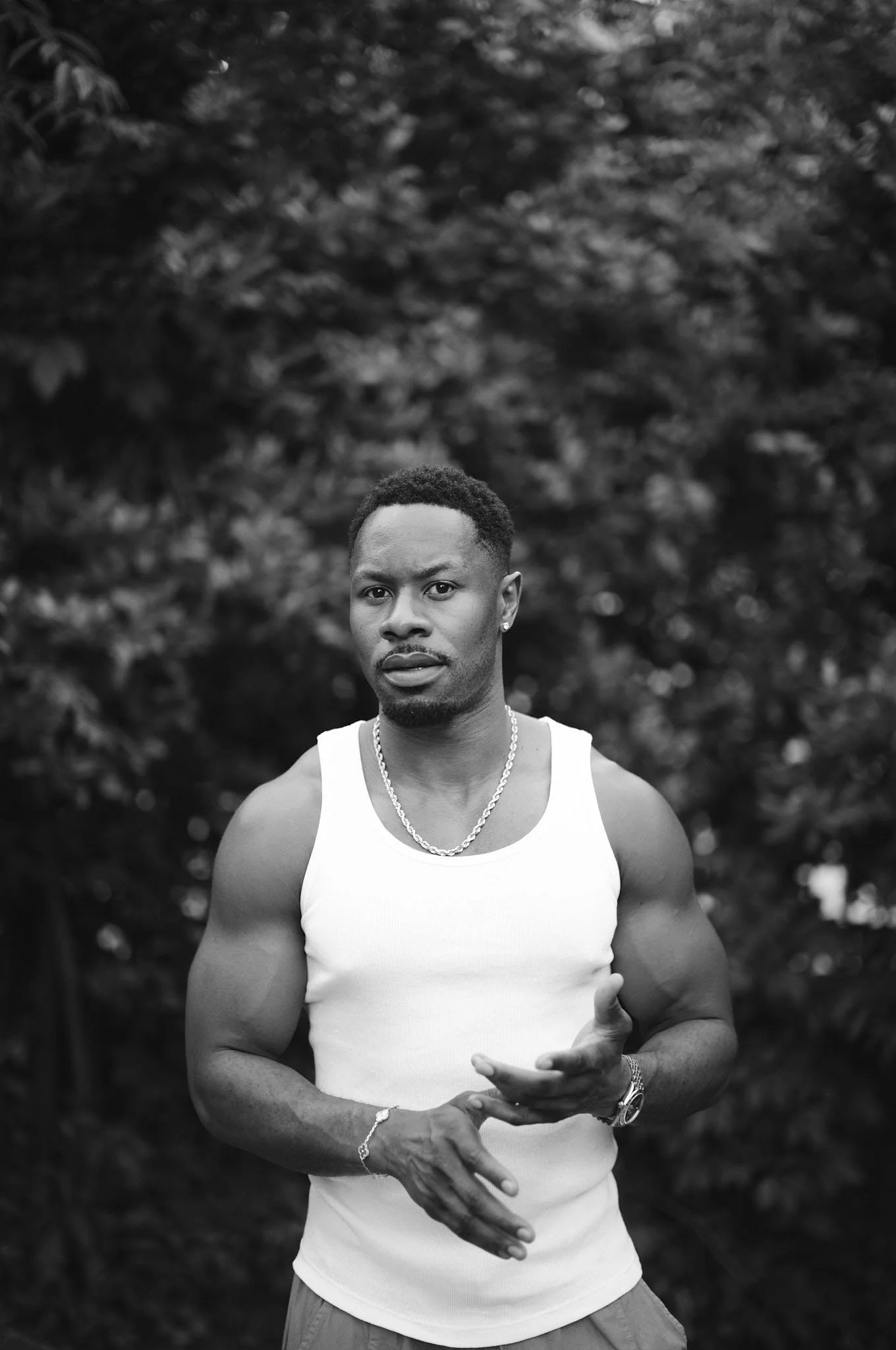 Black and white photo of a young man with short curly hair and a goatee, wearing a sleeveless white shirt, a chain necklace, watch, and bracelet, standing outdoors in front of blurred foliage.