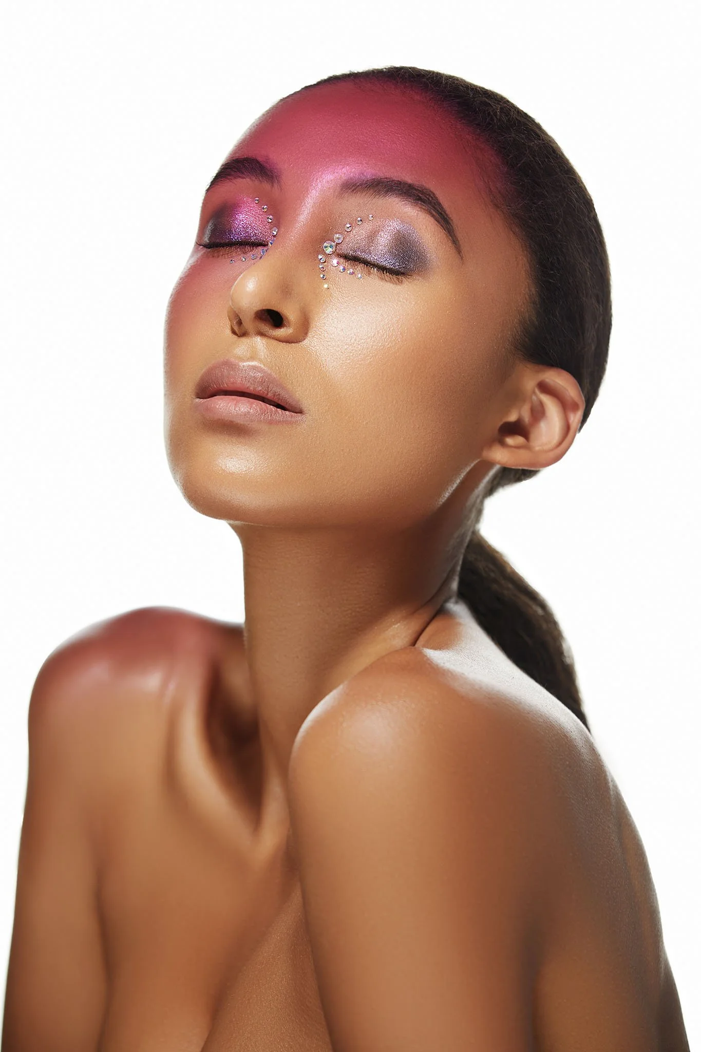 Close-up of a woman with closed eyes, wearing colorful eyeshadow and rhinestone embellishments around her eyes and on her face, with smooth skin and a neutral expression.