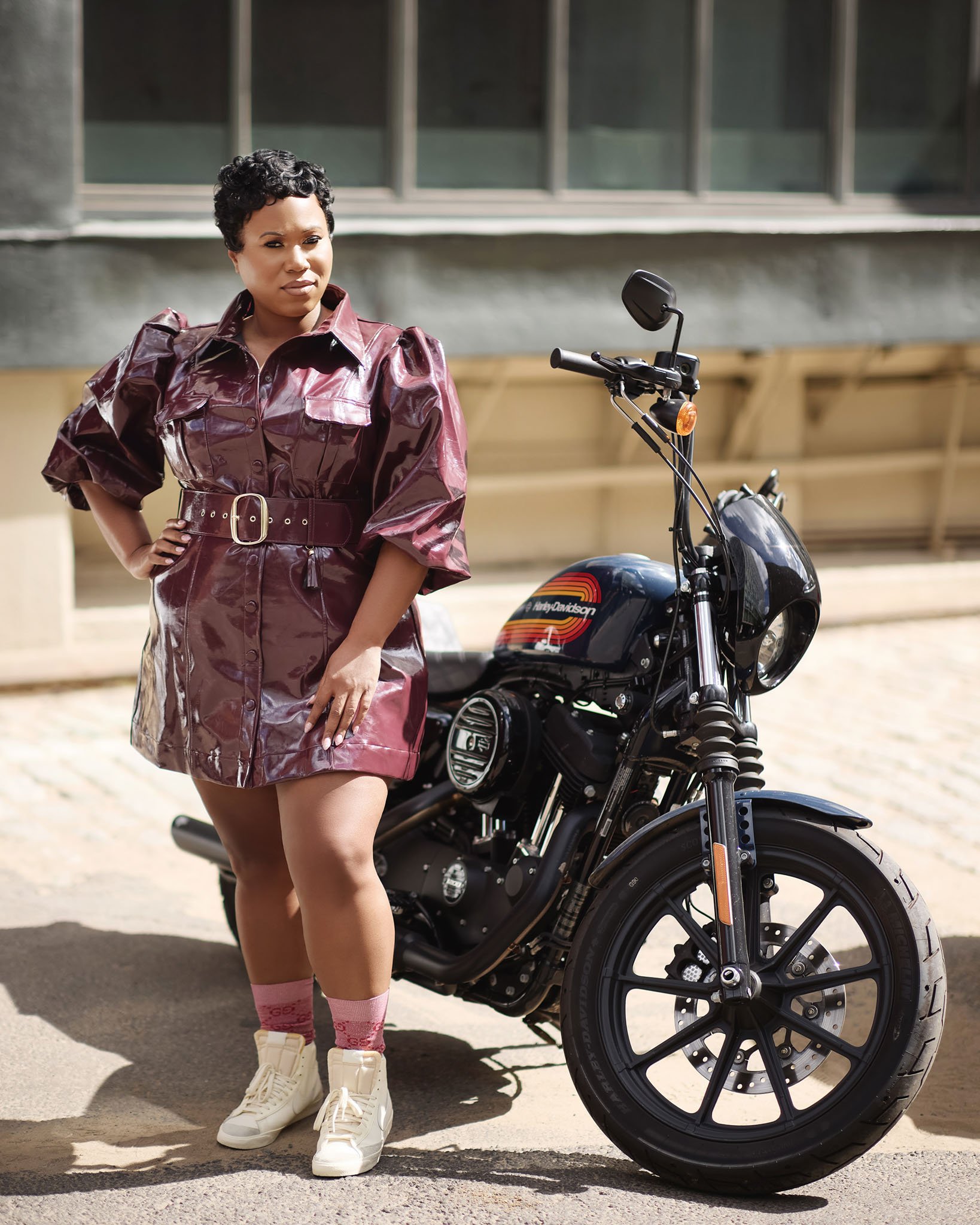 A woman in a shiny burgundy dress with puffed sleeves and a belt stands next to a black Harley Davidson motorcycle on a city street.