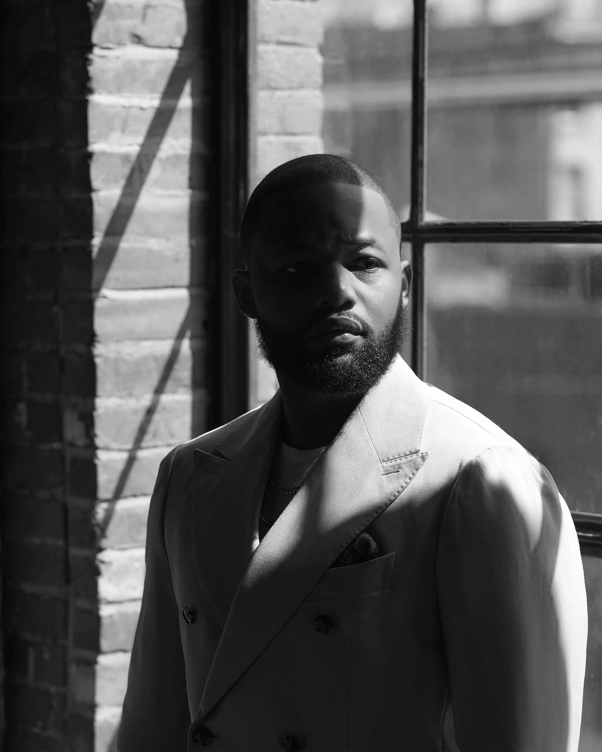 A black and white photo of a man with a beard and short hair, wearing a light-colored blazer, standing near a window with sunlight casting shadows on his face, against a brick wall background.