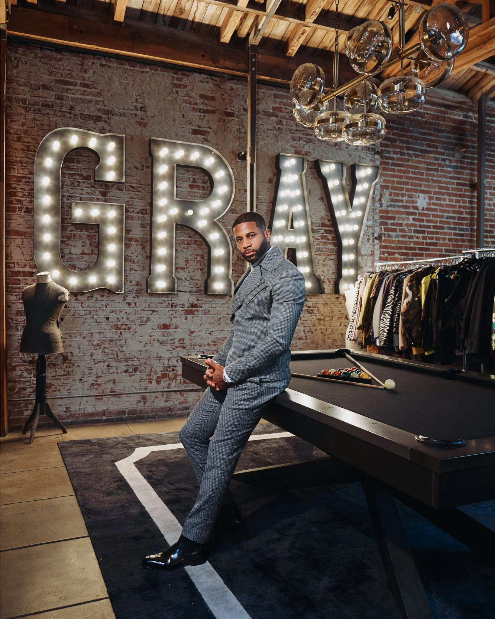 Man in a gray suit sitting at a pool table in a trendy clothing store with a large "GRA Y" sign made of lights on the brick wall behind him, mannequin torso and clothing rack nearby.