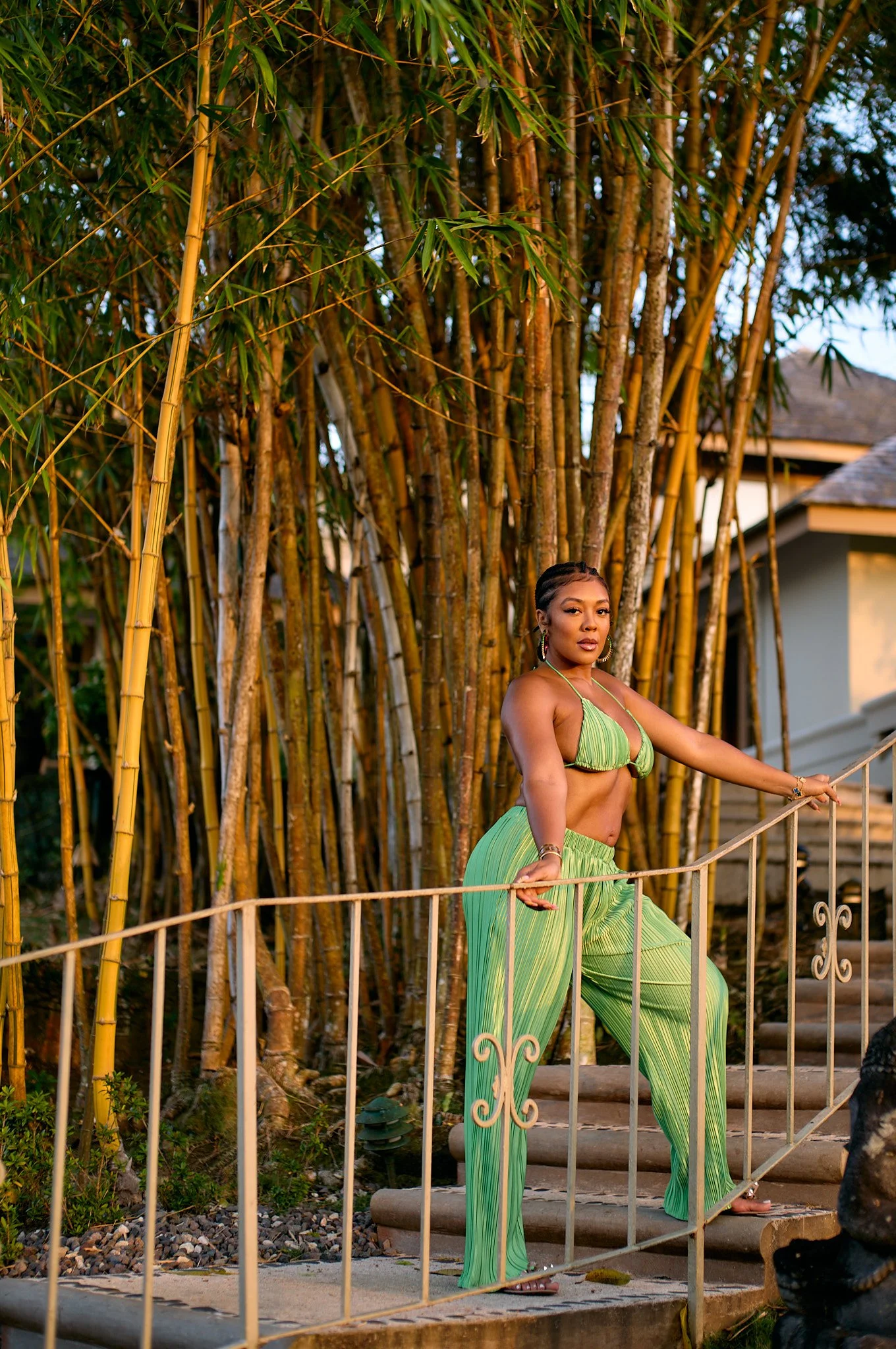 Khadeen Ellis in Jamaica, in green-striped pants and a matching top standing on a staircase outdoors, holding onto a metal railing in front of a backdrop of tall bamboo plants.