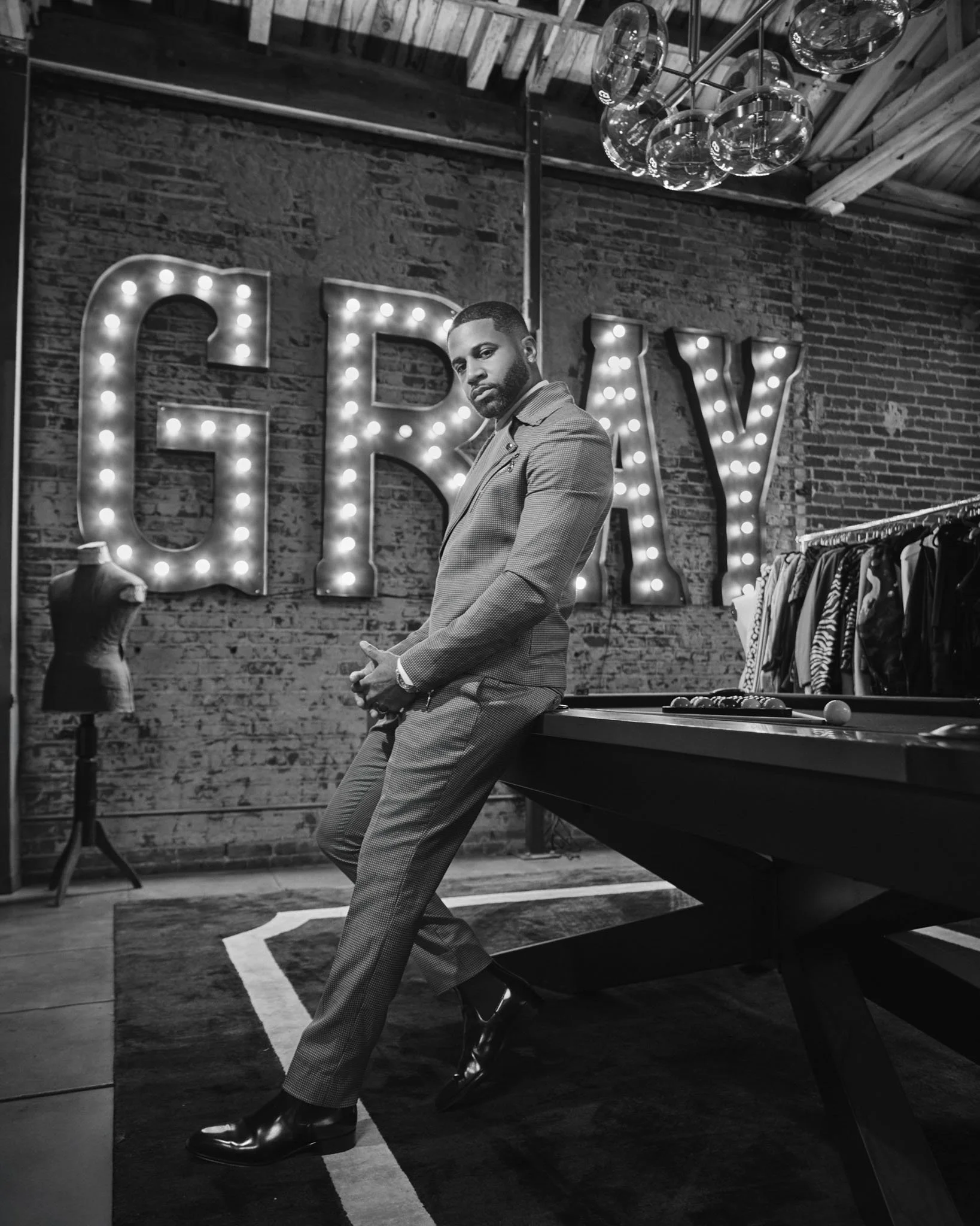 A man in a suit leaning against a pool table in a clothing store with a large illuminated "GIRL" sign on the brick wall behind him.