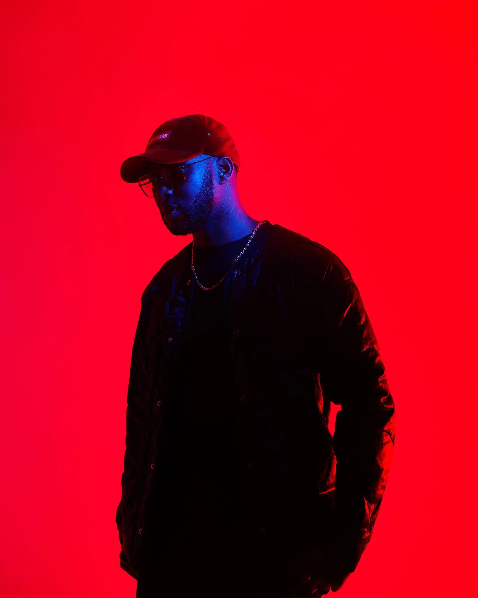 A man wearing sunglasses, a black cap, black shirt, and a shiny black jacket, standing against a vibrant red background.