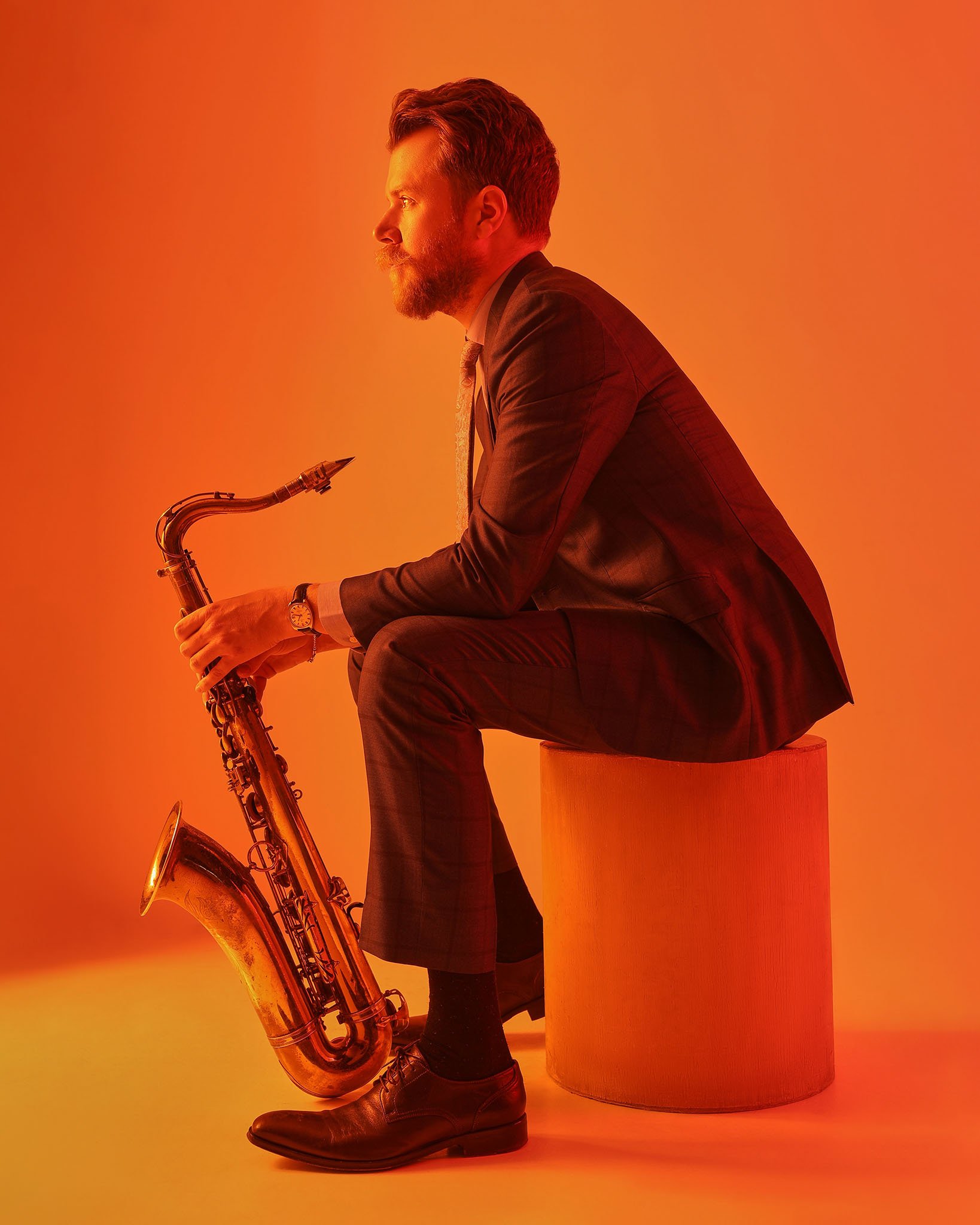 A man in a suit sits on a cylindrical stool, holding a saxophone, with an orange background.