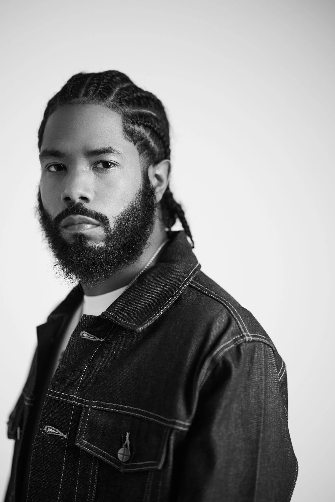 Black and white portrait of a man with braided hair and a full beard, wearing a denim jacket, against a plain light background.