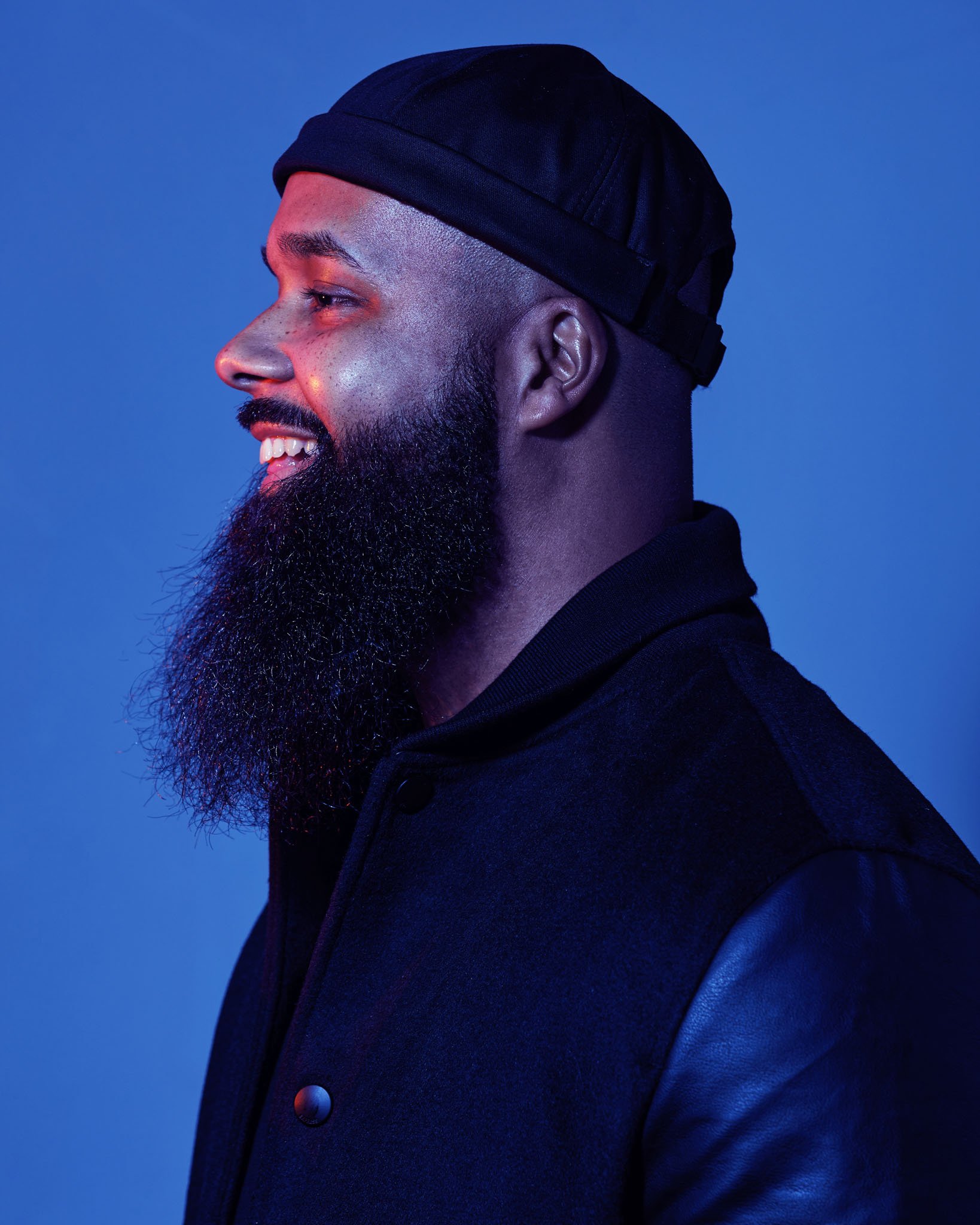 Profile of a smiling man with a beard wearing a black cap and jacket against a blue background.