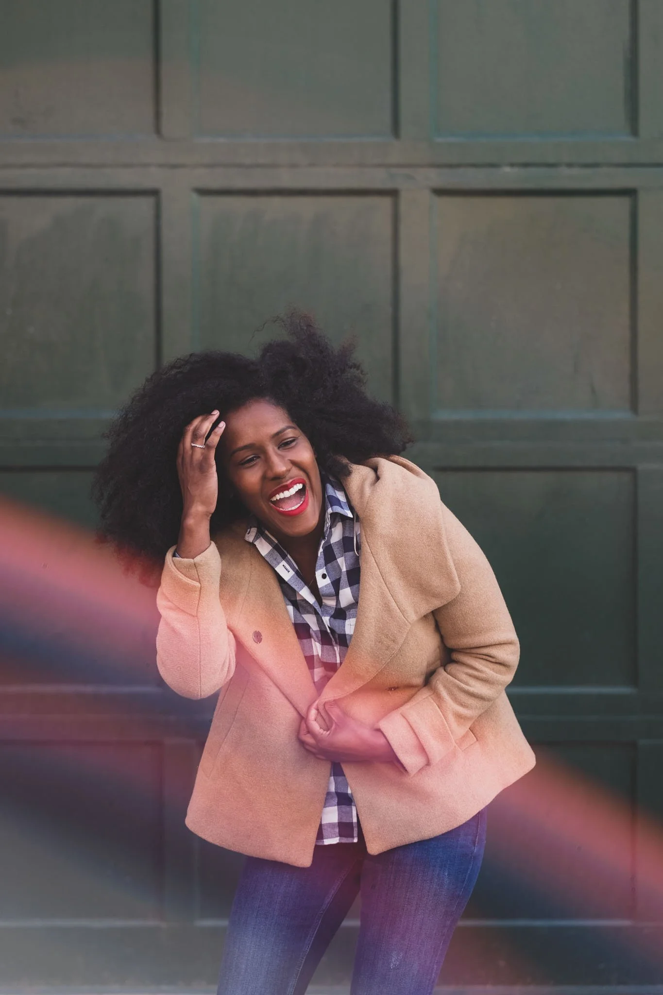 A woman with curly black hair, wearing a tan coat over a plaid shirt, laughing and holding her head with one hand in front of a green garage door.