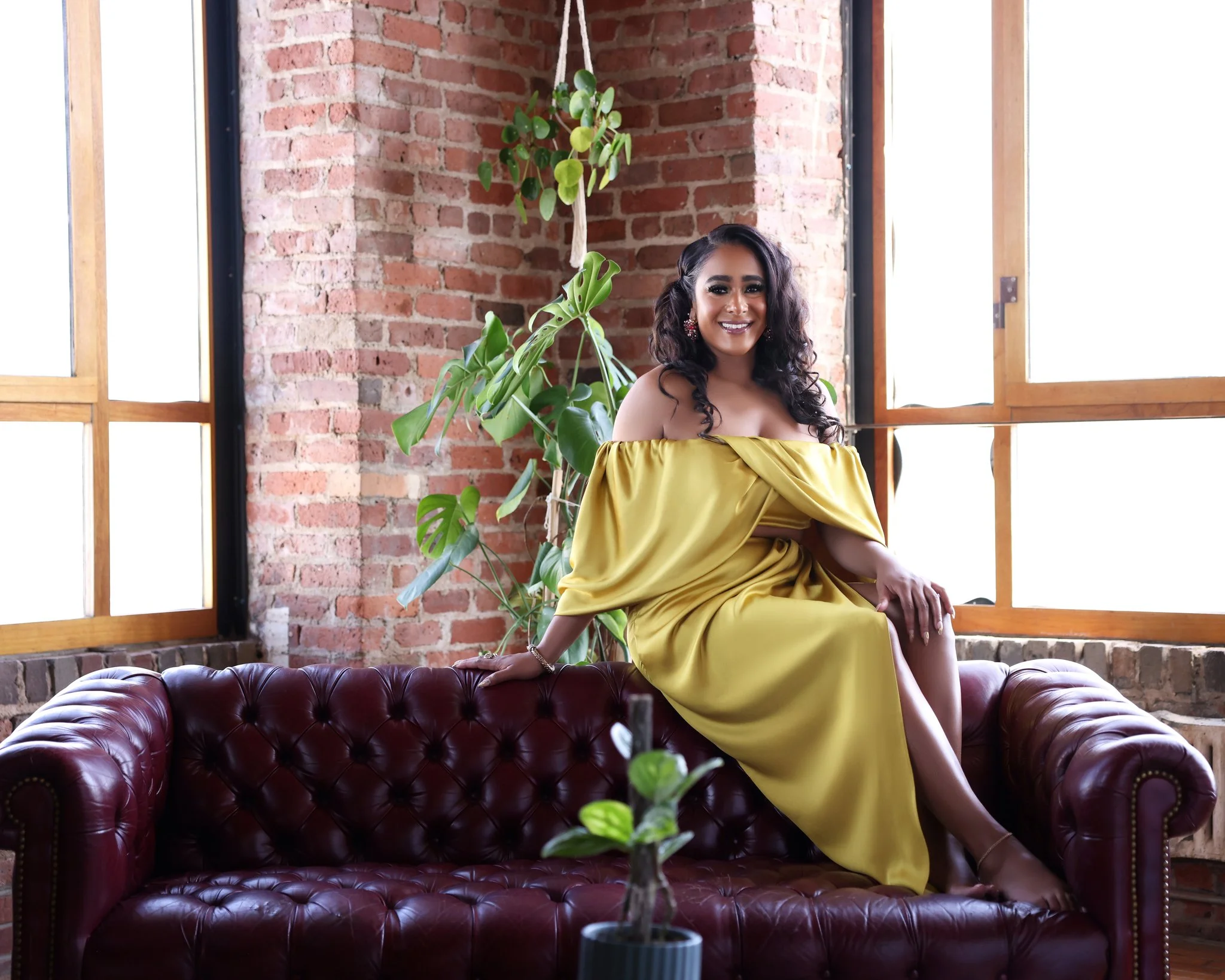 A woman in a yellow off-shoulder dress sitting on a dark red leather chesterfield sofa in a room with exposed brick walls and large windows, smiling at the camera with a green plant behind her.