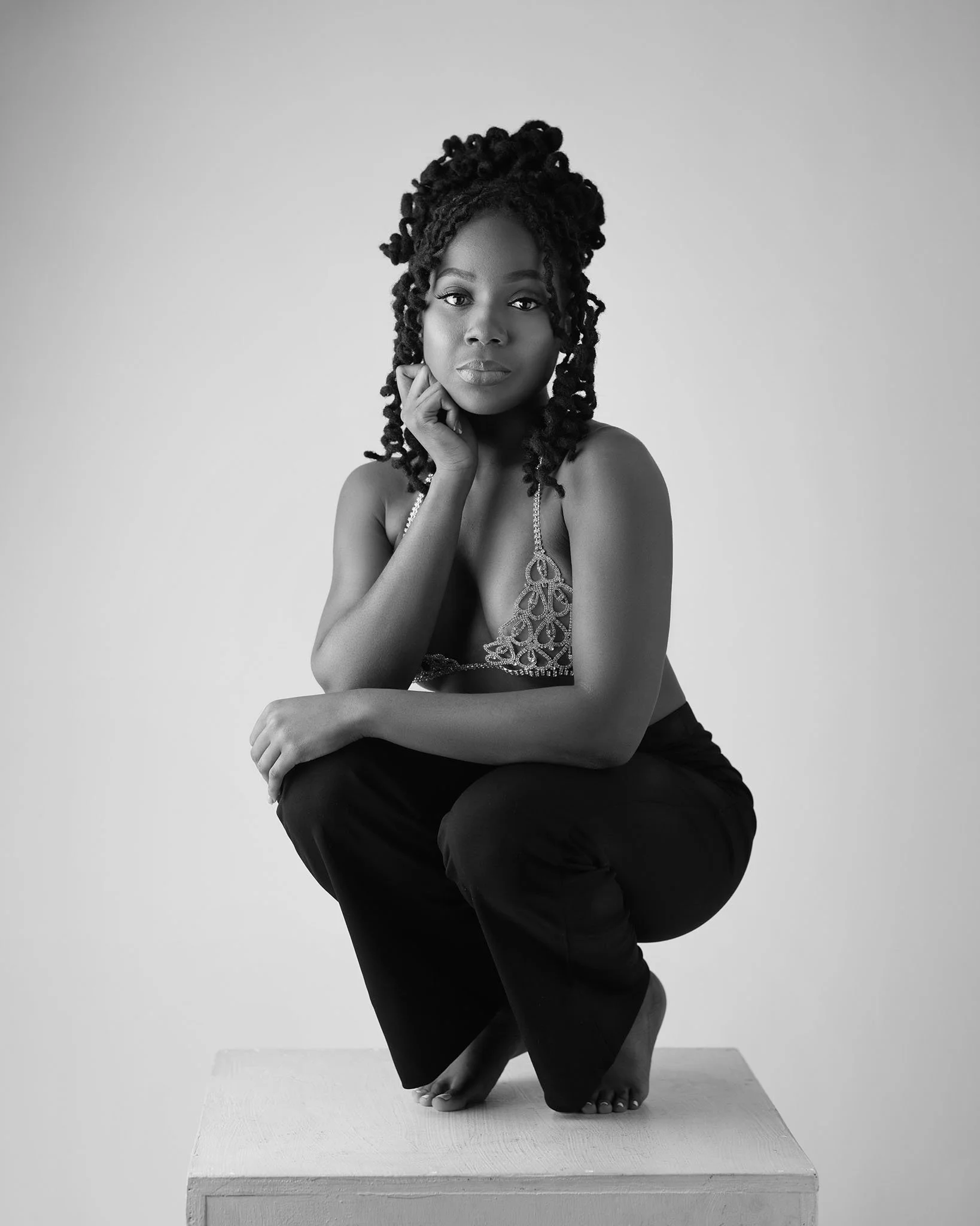 Black and white photo of a woman with long, styled hair, squatting on a platform, wearing a decorative top and black pants, looking at the camera with her chin resting on her hand.