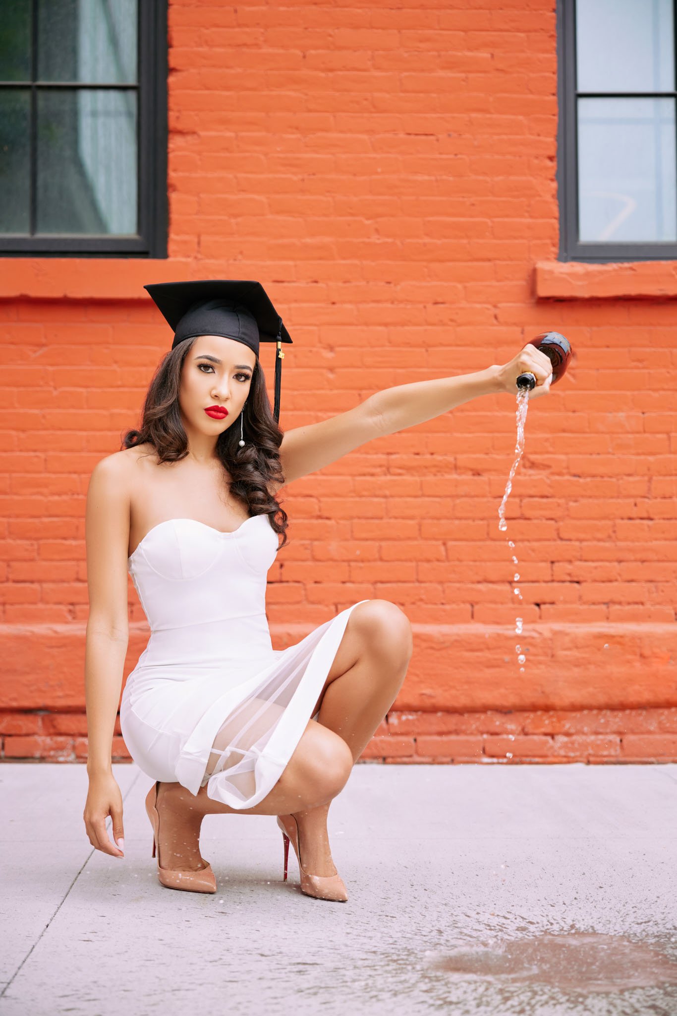 A woman in a white dress and beige high heels crouching on a sidewalk, wearing a black graduation cap, pouring liquid from a bottle onto the ground in front of a red brick wall.