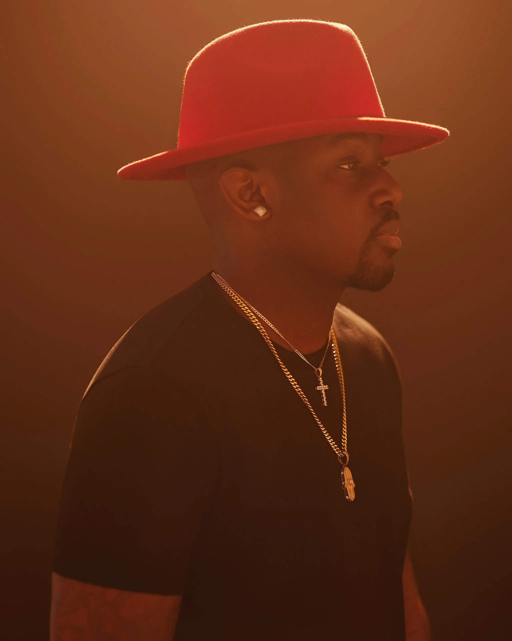 A man wearing a red hat, black shirt, and layered gold jewelry, posed in warm lighting.