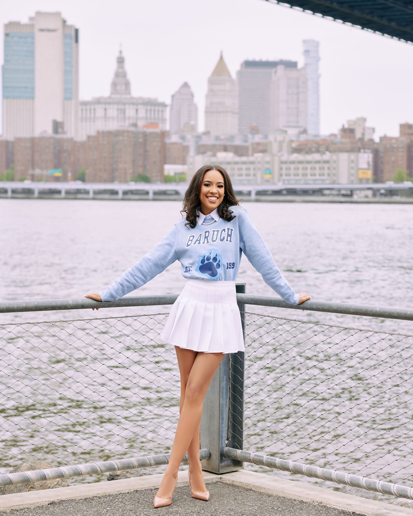 A young woman smiling, wearing a blue sweater with 'Baruch' and a paw print design, a white pleated skirt, and nude heels, standing by a riverfront with a city skyline in the background.