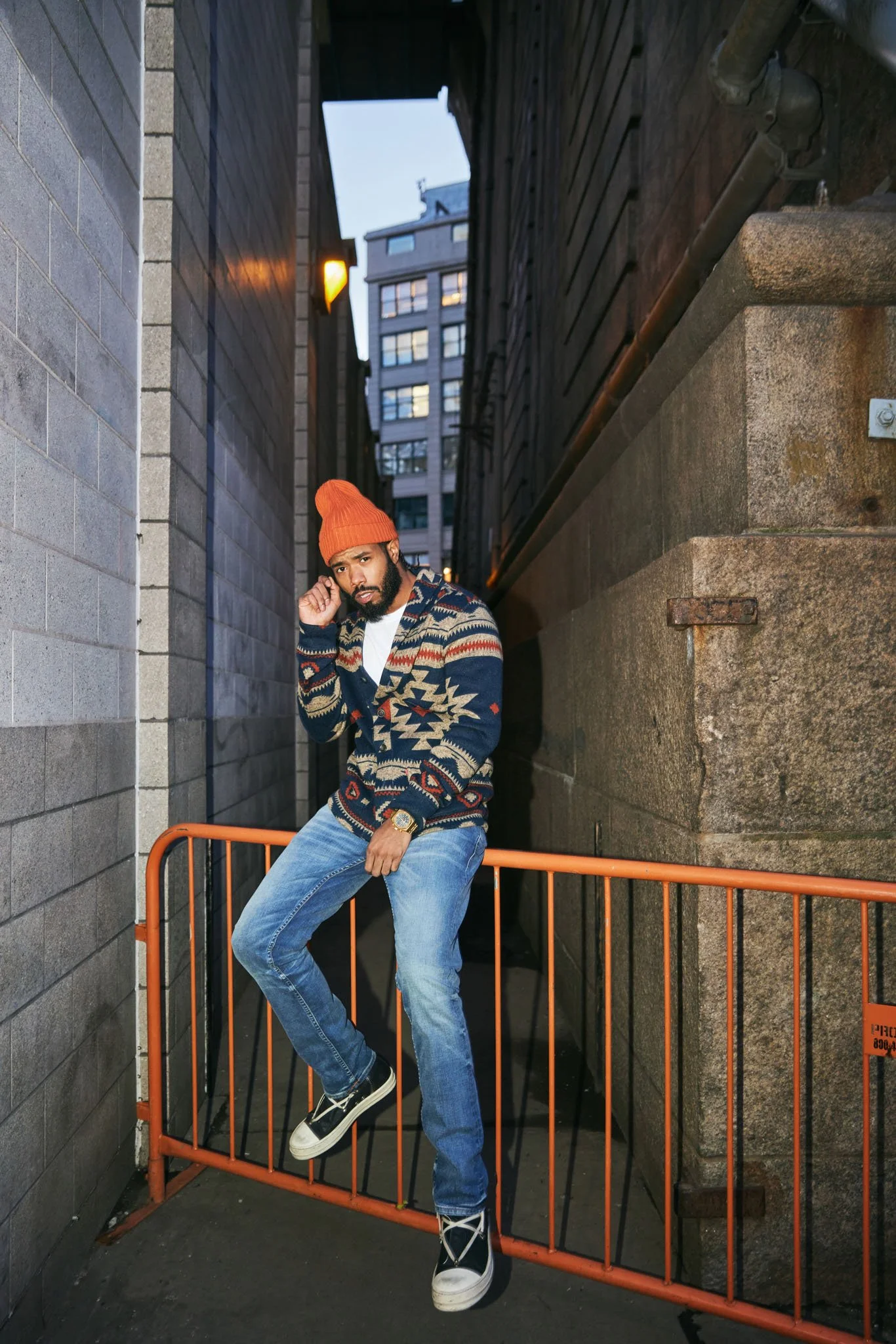 A man wearing an orange beanie, patterned sweater, blue jeans, and sneakers, leaning on an orange metal barricade in an urban alleyway during evening.