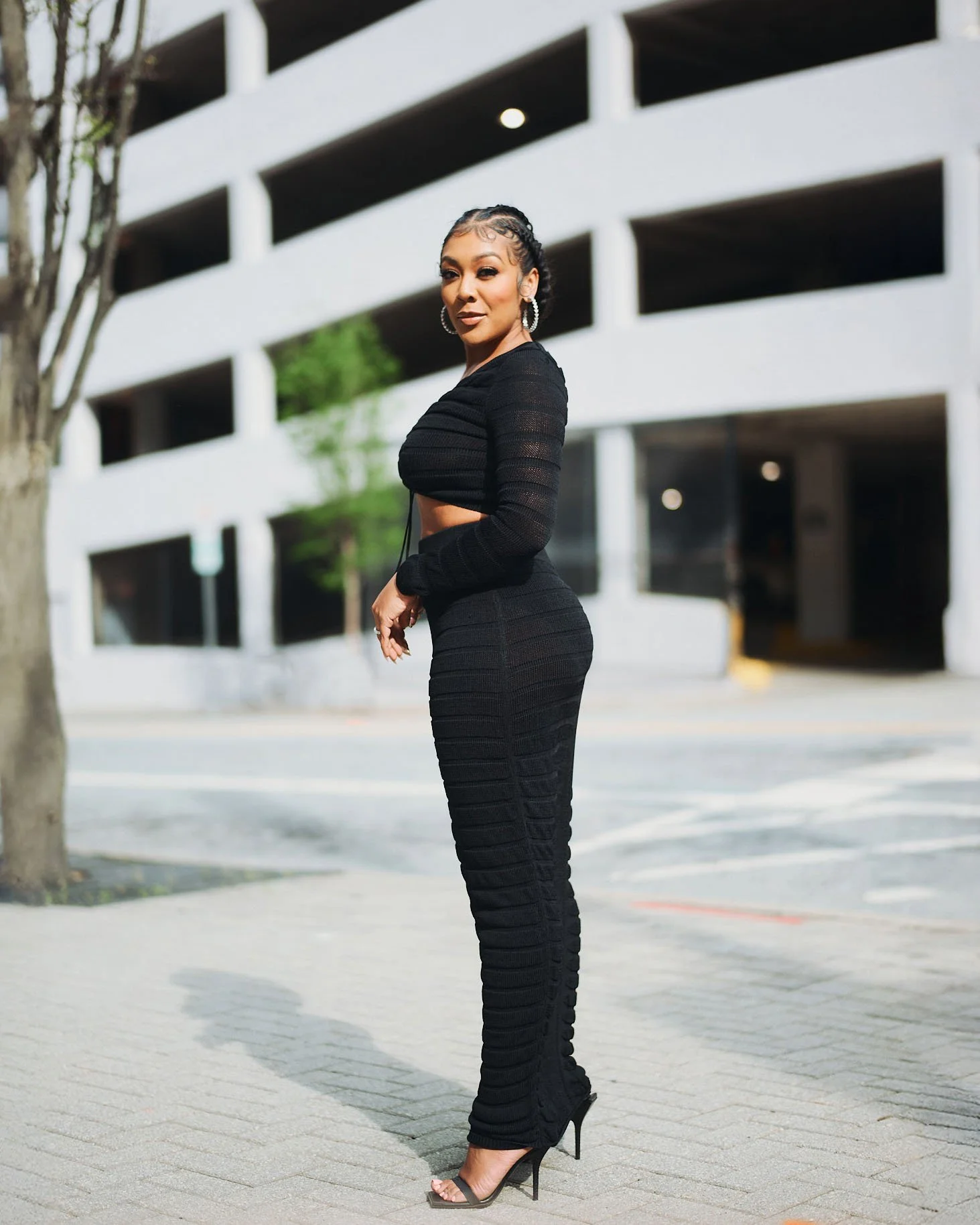 A woman dressed in a black, long-sleeve, crop top and matching high-waisted pants, standing on a sidewalk in an urban area with a multi-story parking garage in the background. She is wearing black high heels and large hoop earrings.