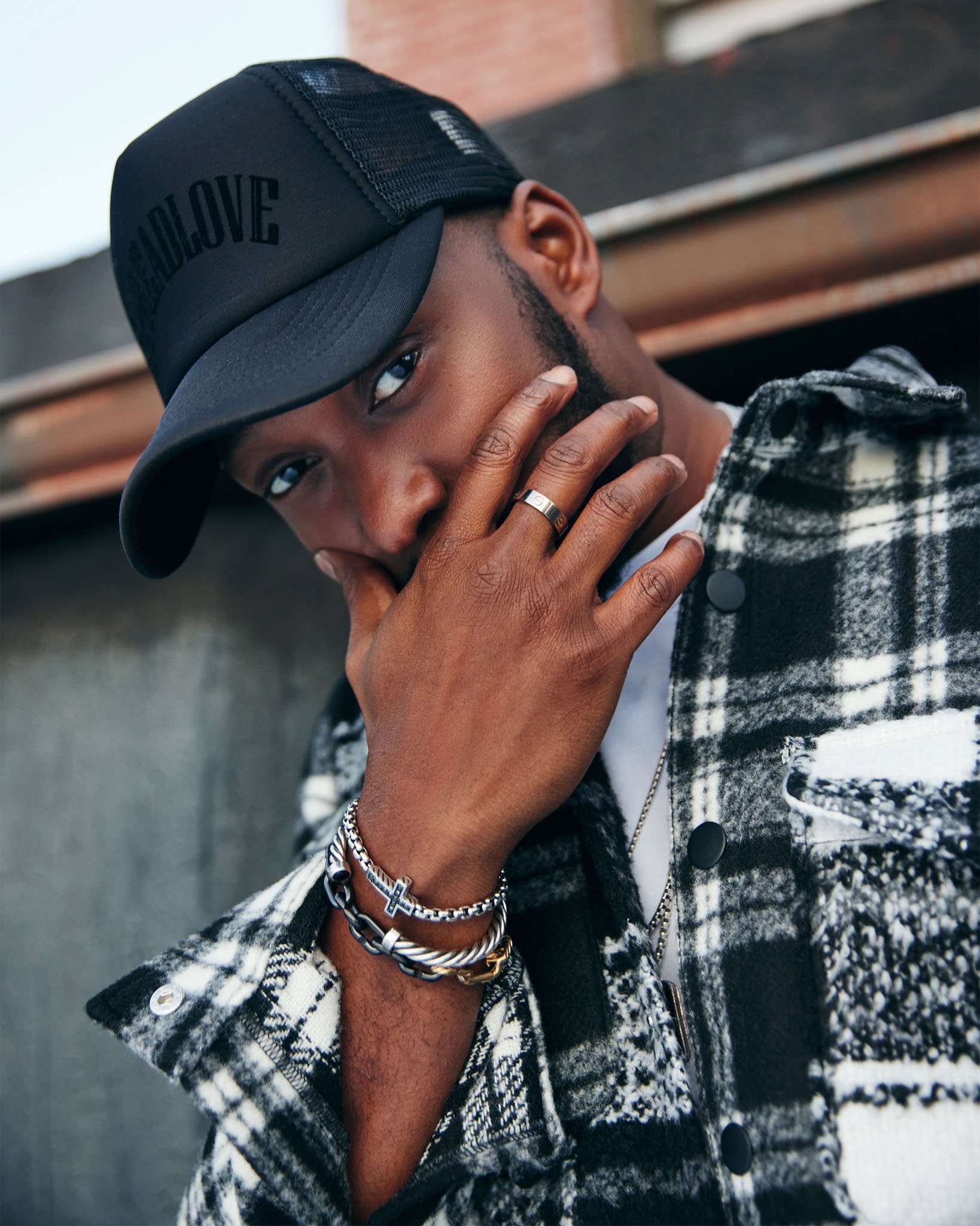 A young man with medium brown skin tone wearing a black cap, a plaid jacket, and layered bracelets, with his hand partially covering his mouth and looking into the camera.