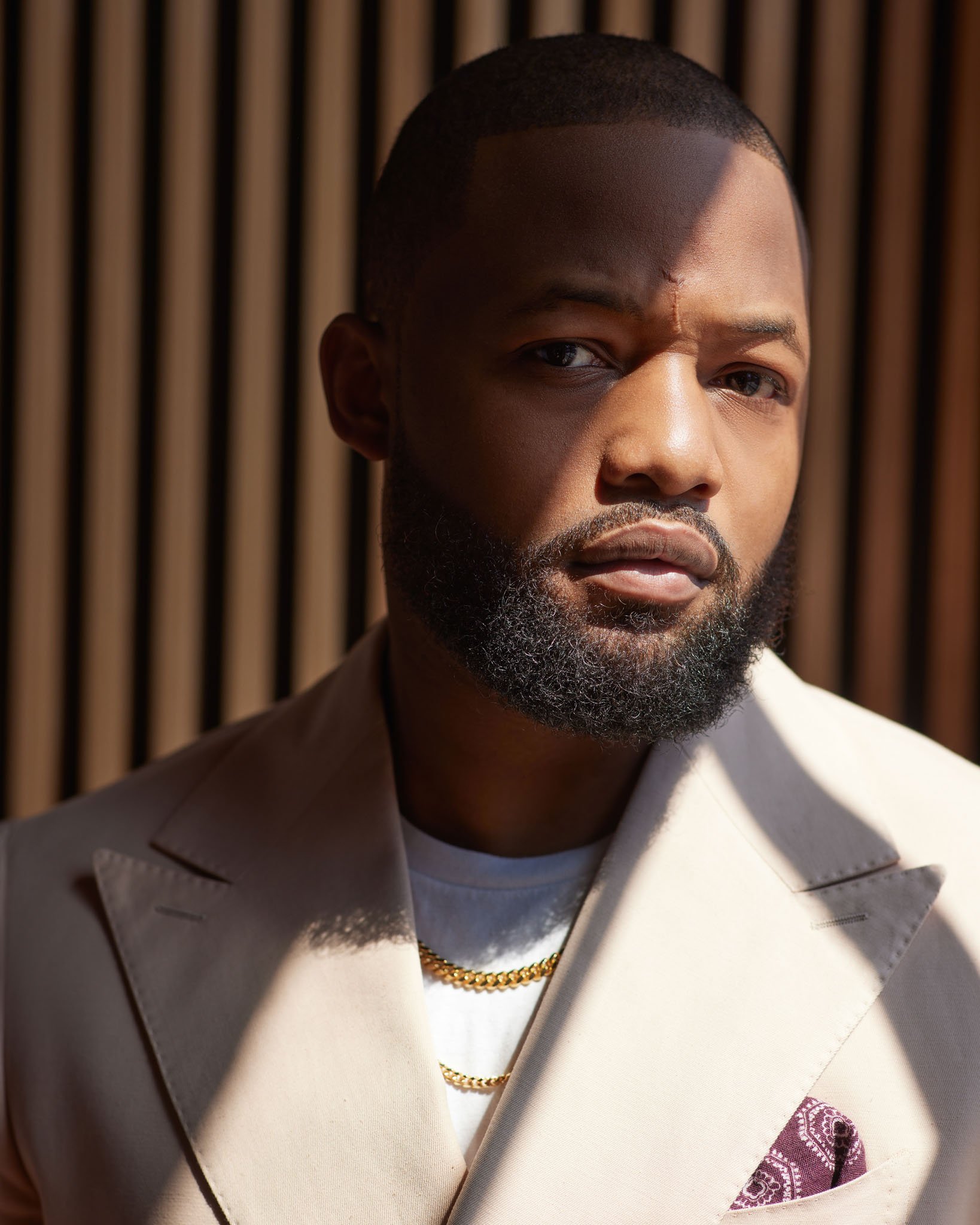 A young man with a beard and short hairstyle, wearing a white suit, gold chain, and a pocket square, standing in front of a wooden background with shadows cast across his face.
