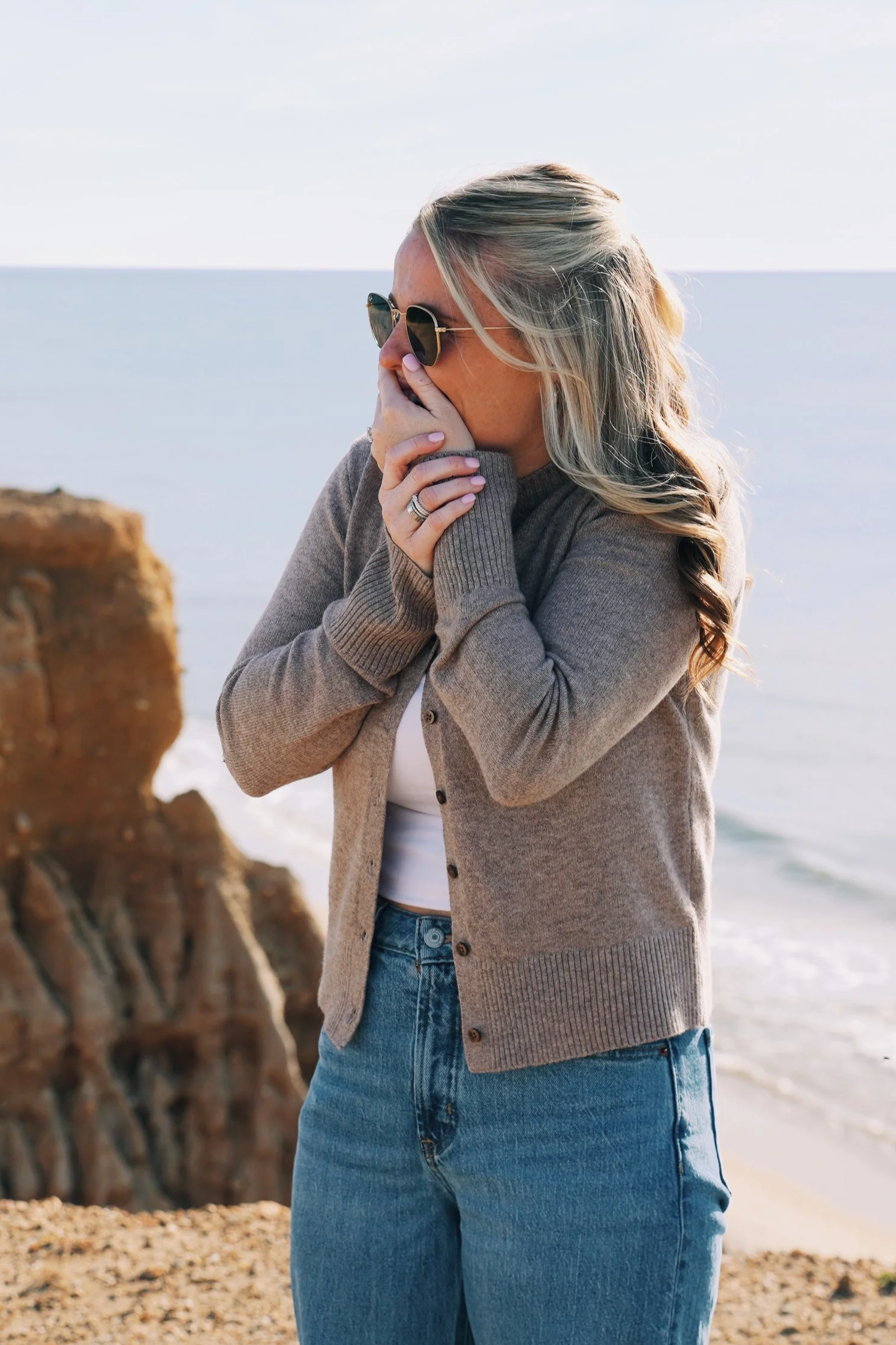 A woman with blonde hair wearing sunglasses, a brown cardigan, and blue jeans, standing near the beach with rocks and ocean in the background.