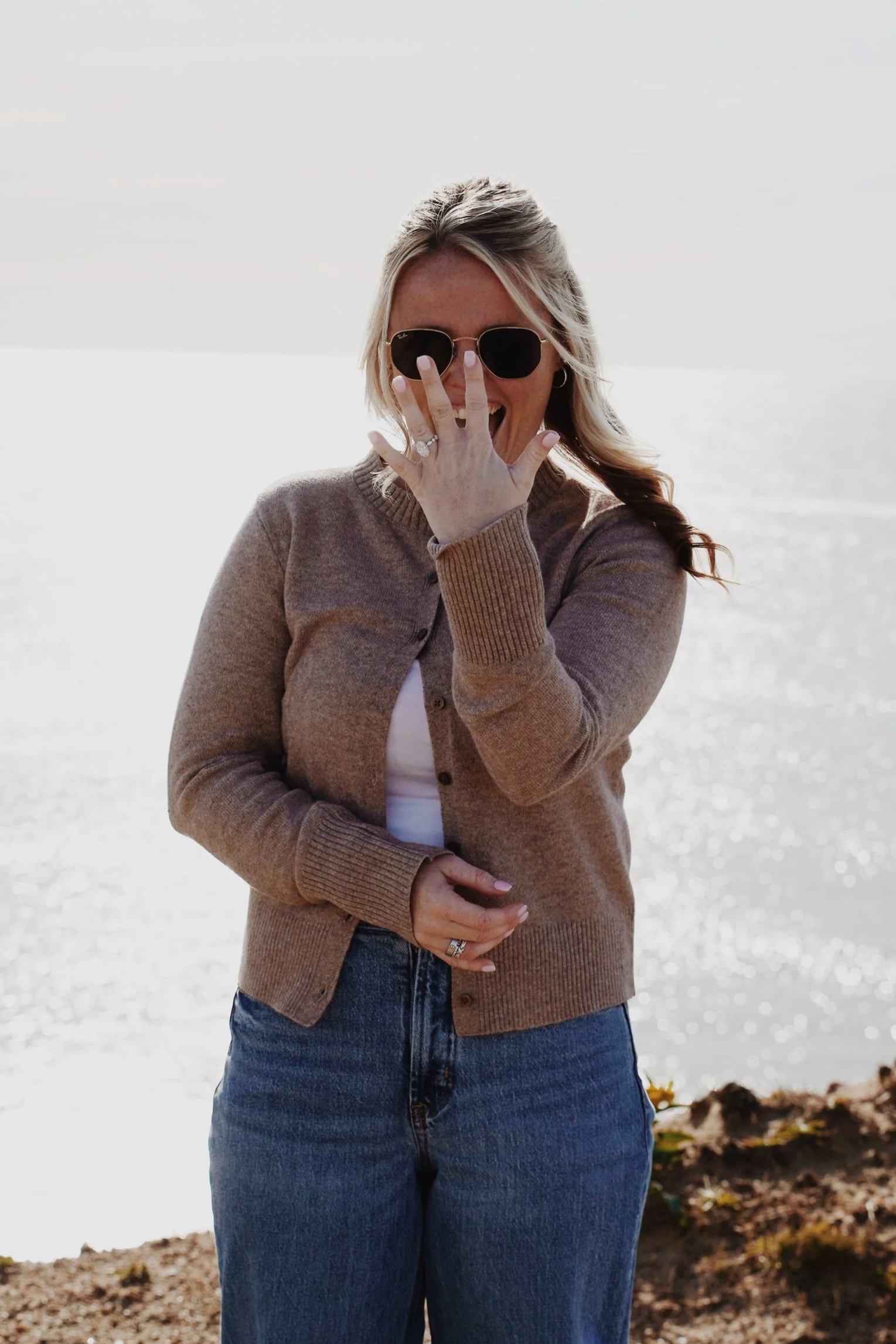 A smiling woman with blond hair, wearing sunglasses, a beige cardigan, and blue jeans, standing on a beach with the ocean in the background.