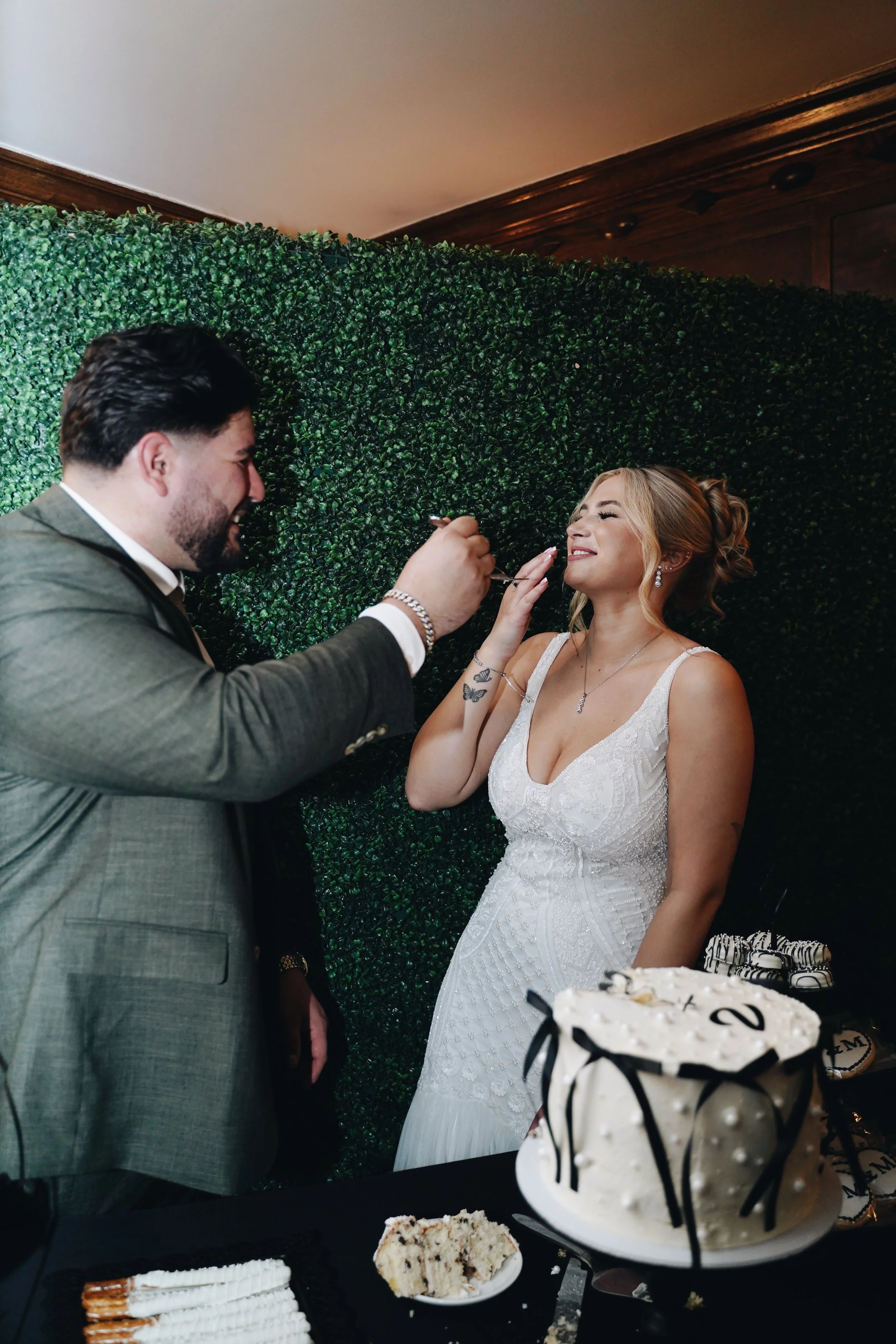 A man is feeding a woman wedding cake at a celebration, with a decorated green hedge background and a table of desserts in the foreground.