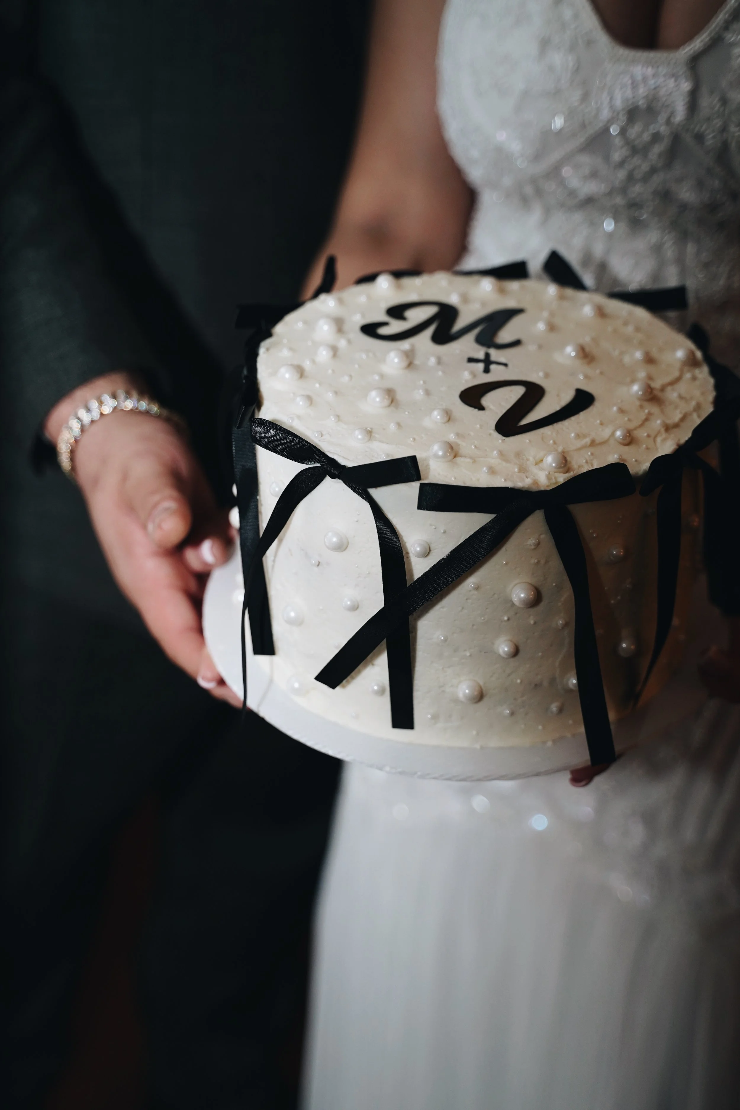 A person wearing a black dress and pearl jewelry holds a white, oval-shaped cake decorated with pearls, black bows, and black monogram initials 'M + V' on top.