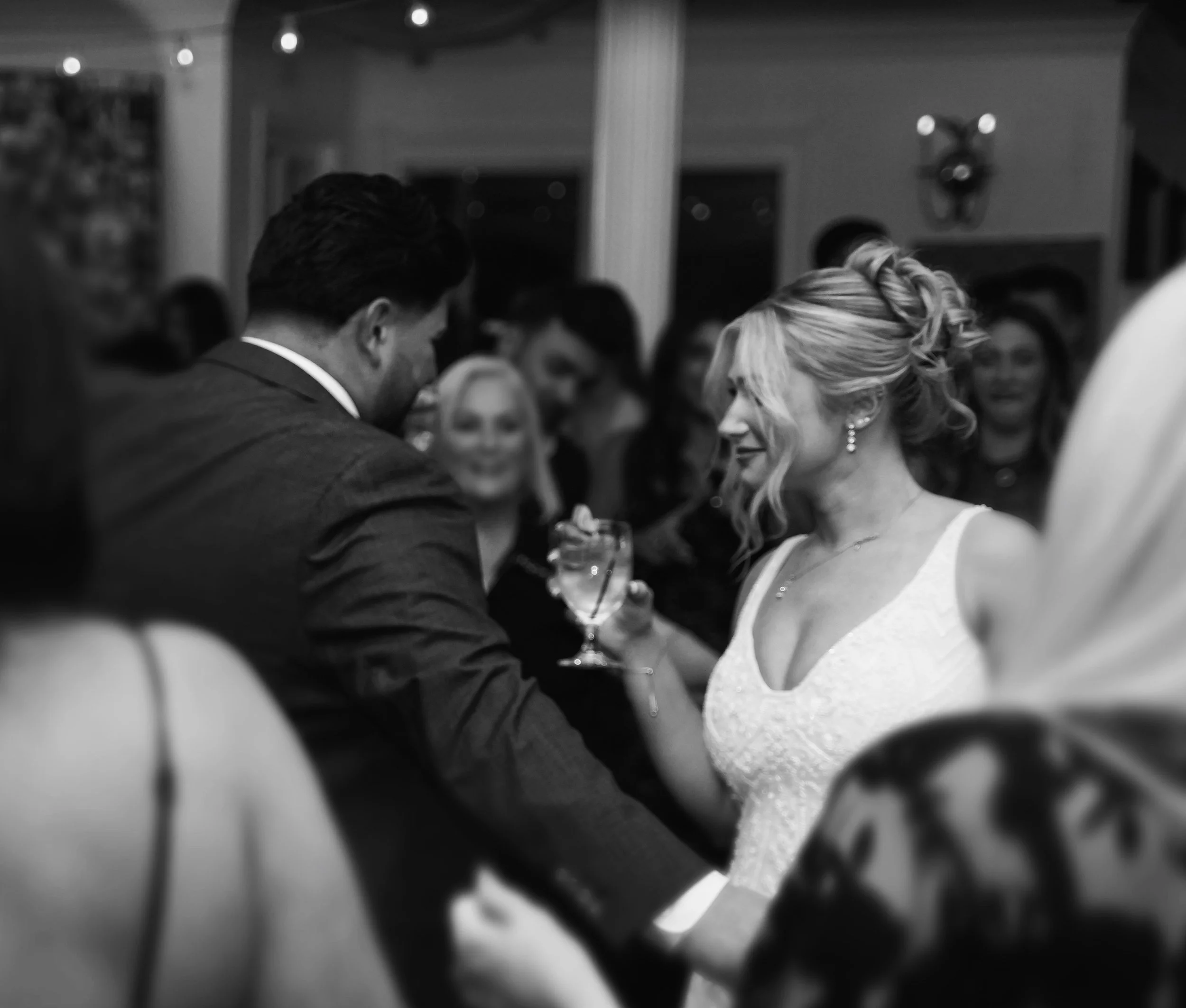 Black and white photo showing a group of people at a social gathering, with some engaged in conversation and holding drinks. The setting appears to be indoors with warm lighting.