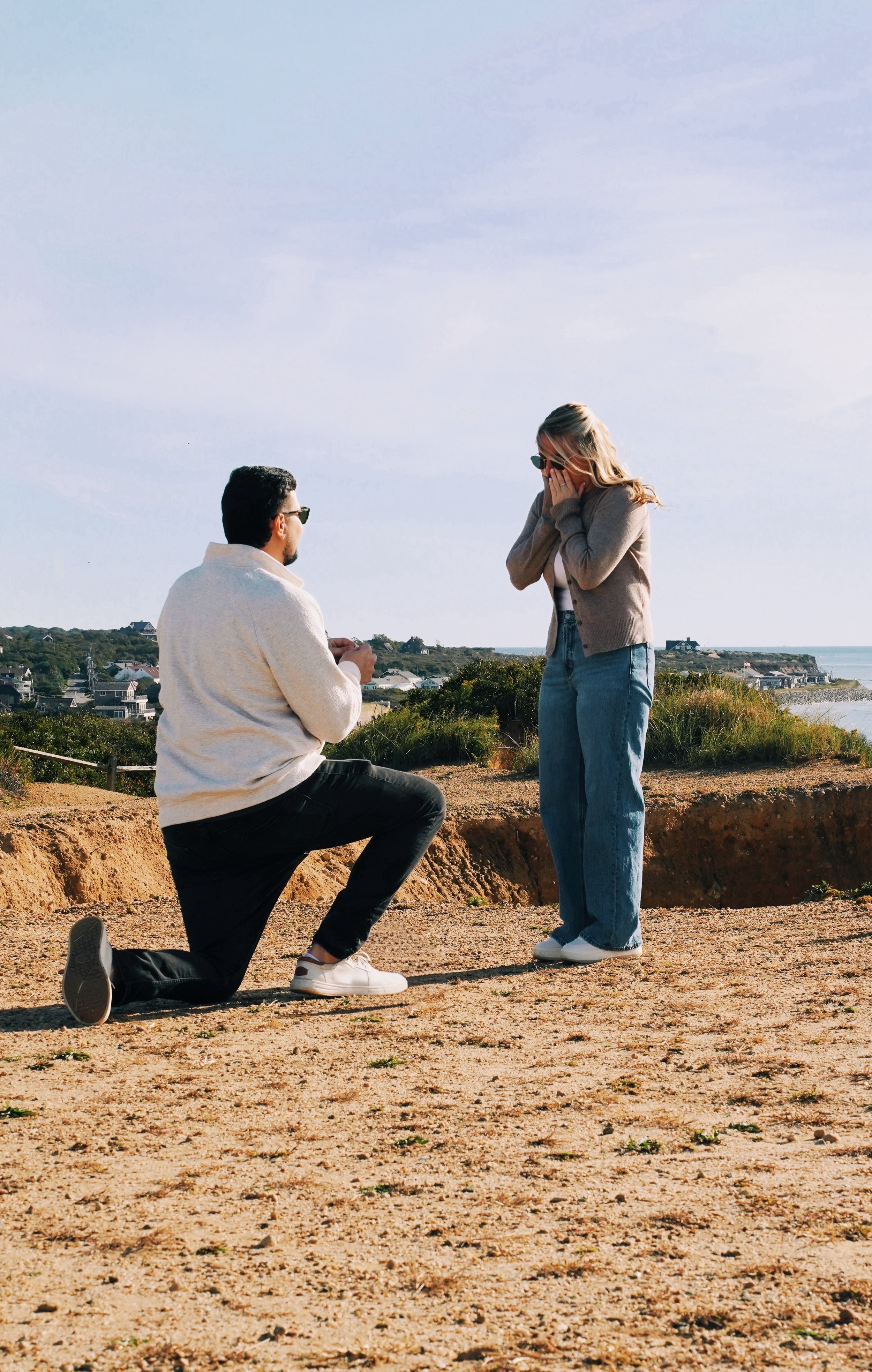 A man kneeling on one knee proposing to a woman outdoors on a sandy area with houses and ocean in the background.