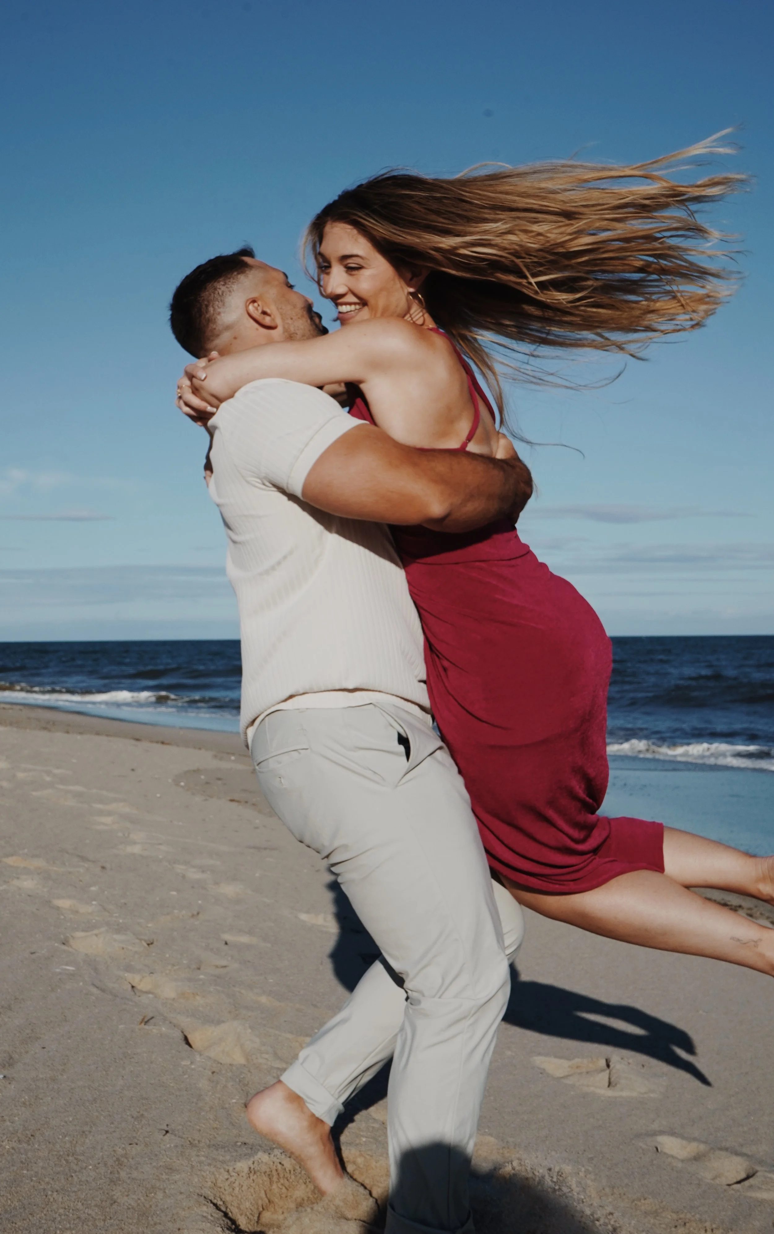 A man lifting a woman at the beach. The woman has long hair flowing in the wind, and they are smiling while looking at each other. The sandy shoreline and ocean are in the background.