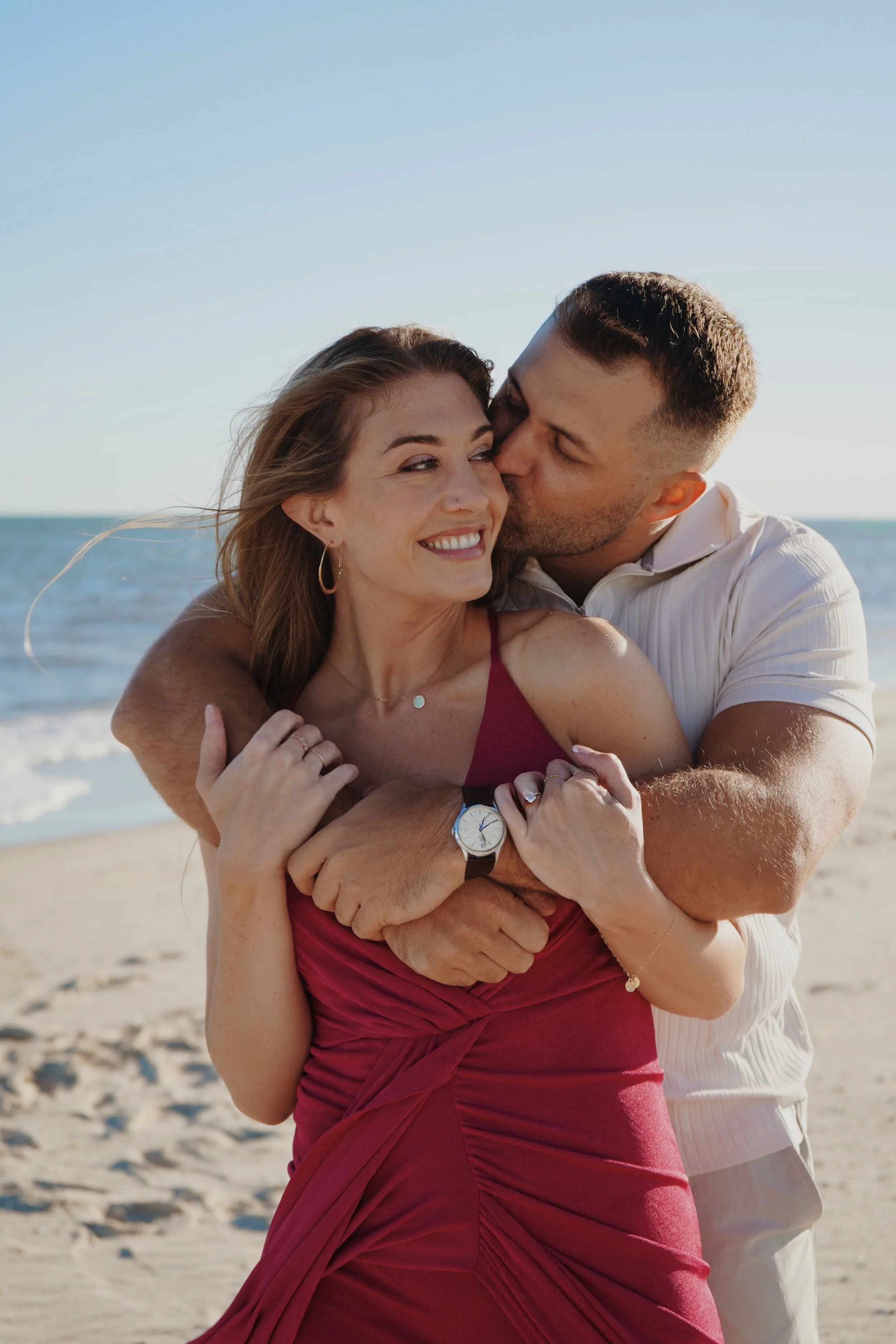 A couple embraces and shares a kiss at the beach, with the woman smiling and the man kissing her cheek, against a backdrop of sandy shore and ocean water.