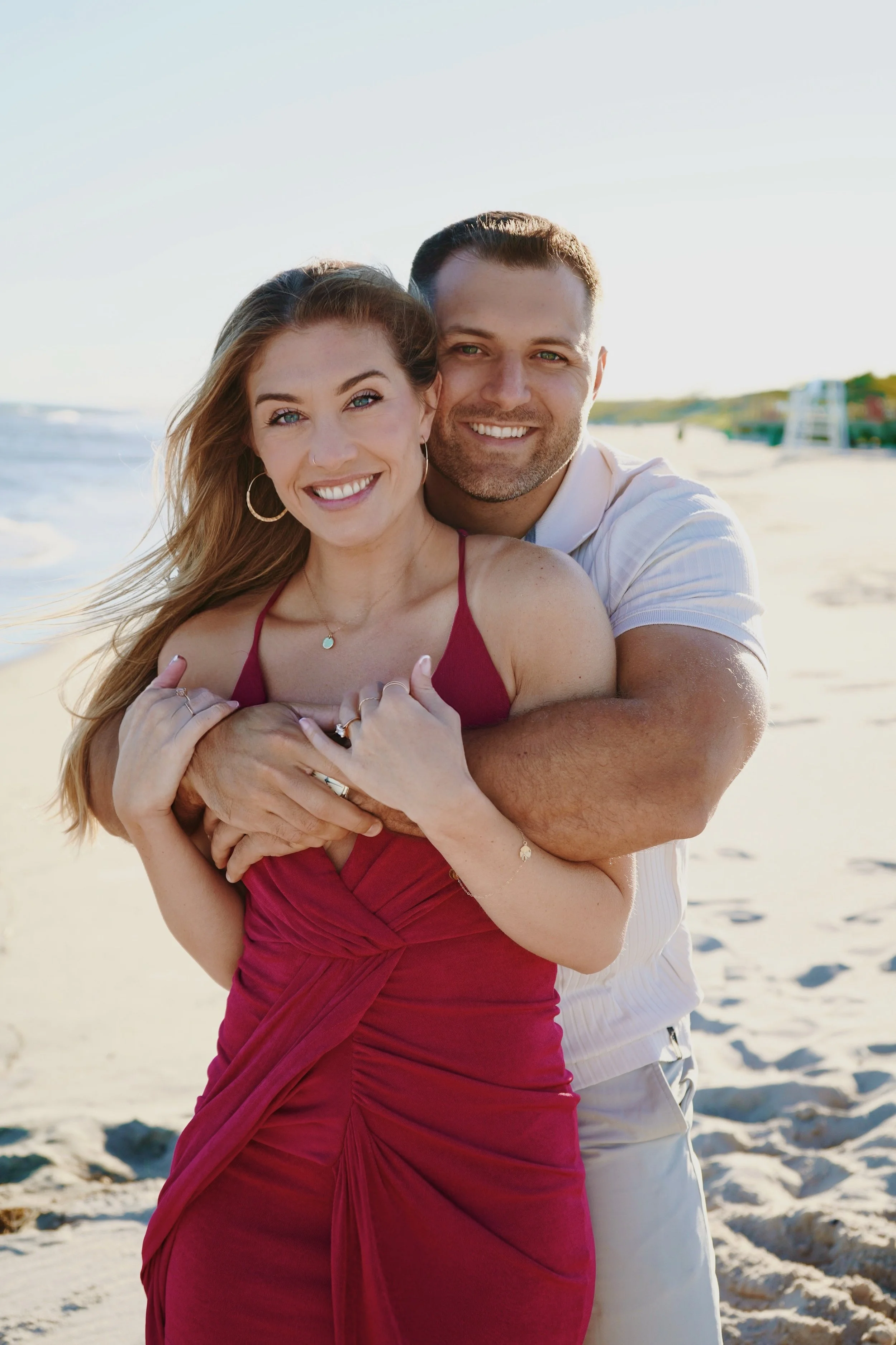 A smiling couple embracing on a beach, with the woman in a red dress and the man in a white shirt, sunset in the background.