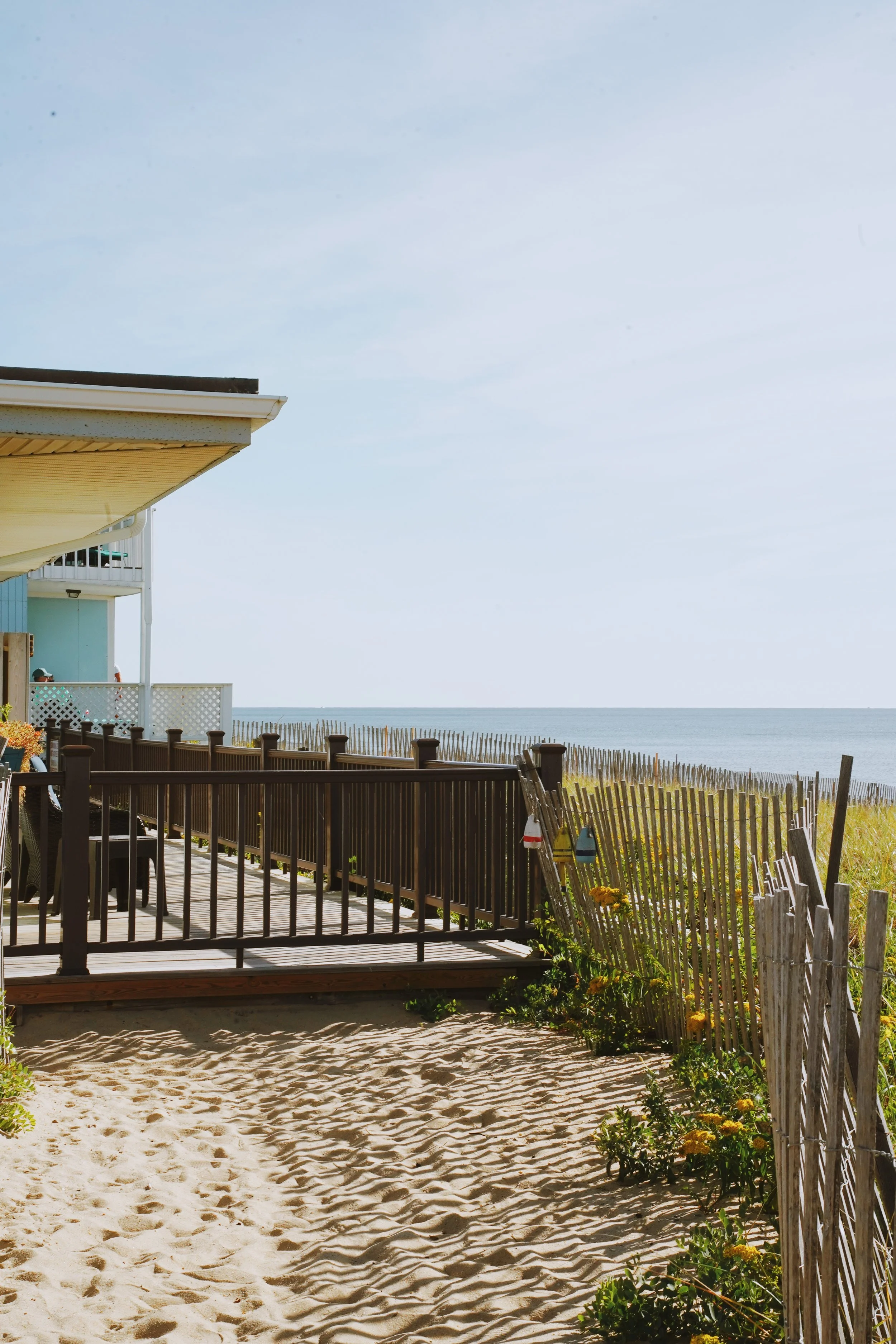 Beach scene with sandy shore, wooden fence, ocean in the background, and part of a house with a balcony.