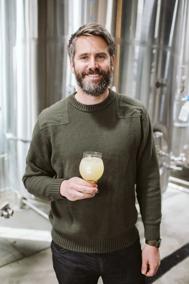 A man with a beard and short hair, wearing a green sweater and black pants, is holding a glass of pale yellow beverage in a brewery with large metal tanks in the background.