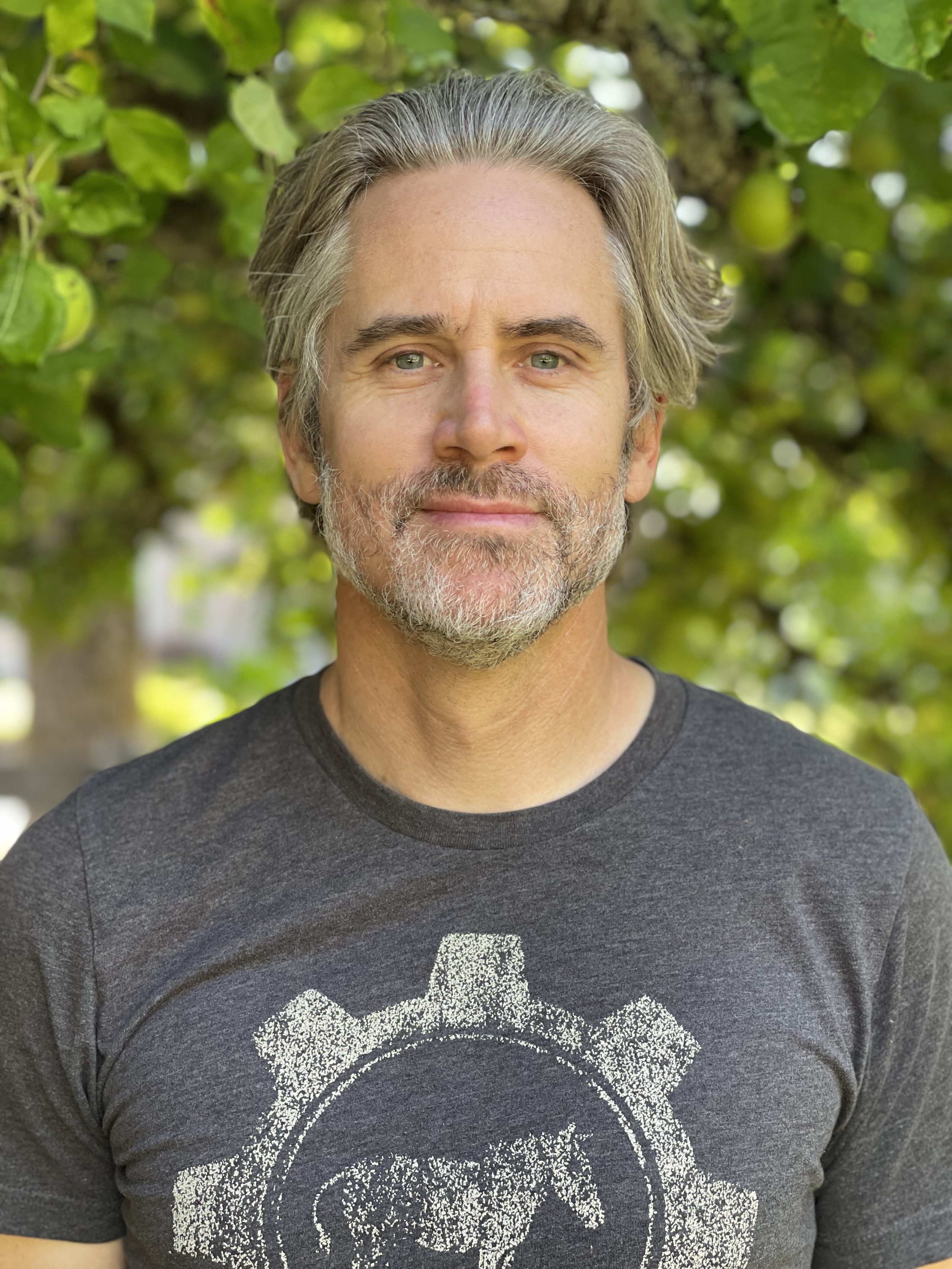 Close-up of a middle-aged man with gray hair and beard, smiling, outdoors with green leaves in the background, wearing a dark gray t-shirt with a glittery skull graphic.